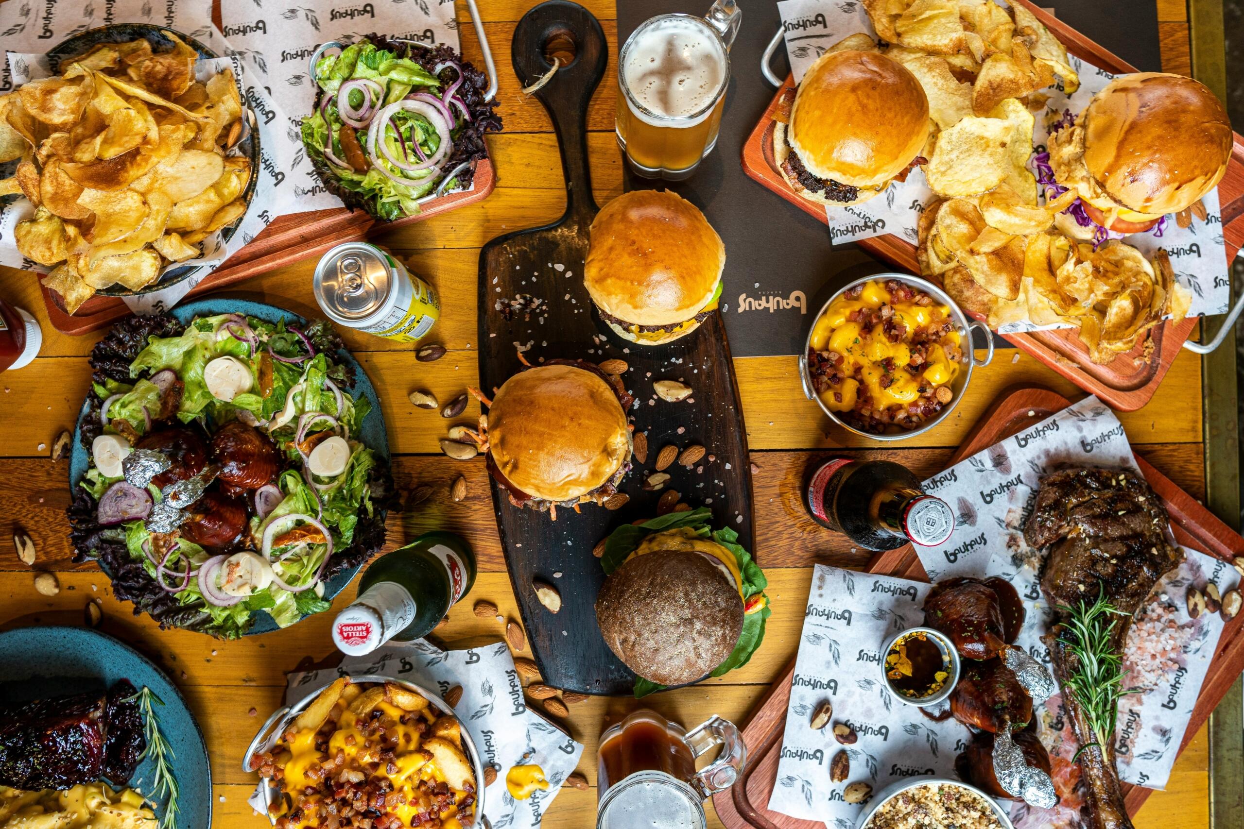 Τραπέζι γεμάτο φαγητά τύπου street food με μπέργκερ, πατάτες, σαλάτες και ποτά σε ξύλινη επιφάνεια.