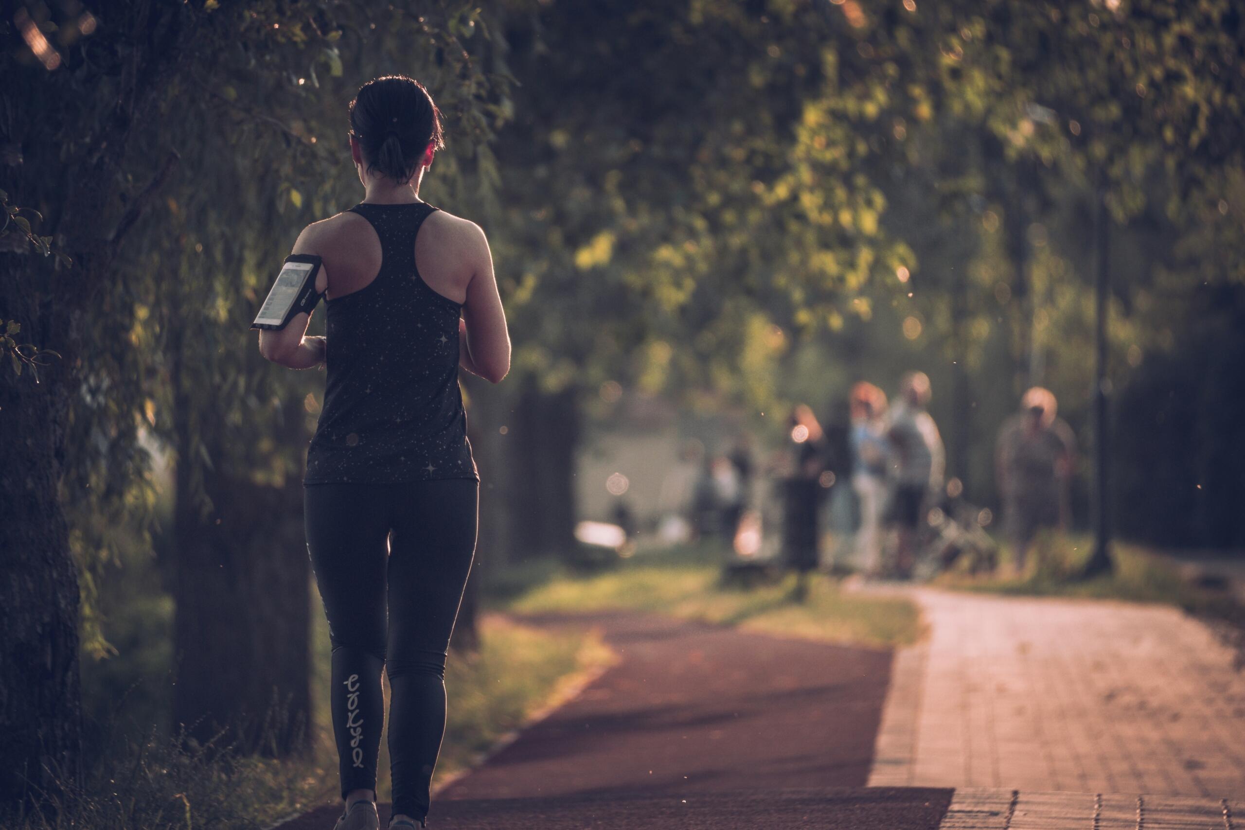 Γυναίκα κάνει jogging σε πάρκο φορώντας αθλητικά ρούχα και θήκη κινητού στο μπράτσο.