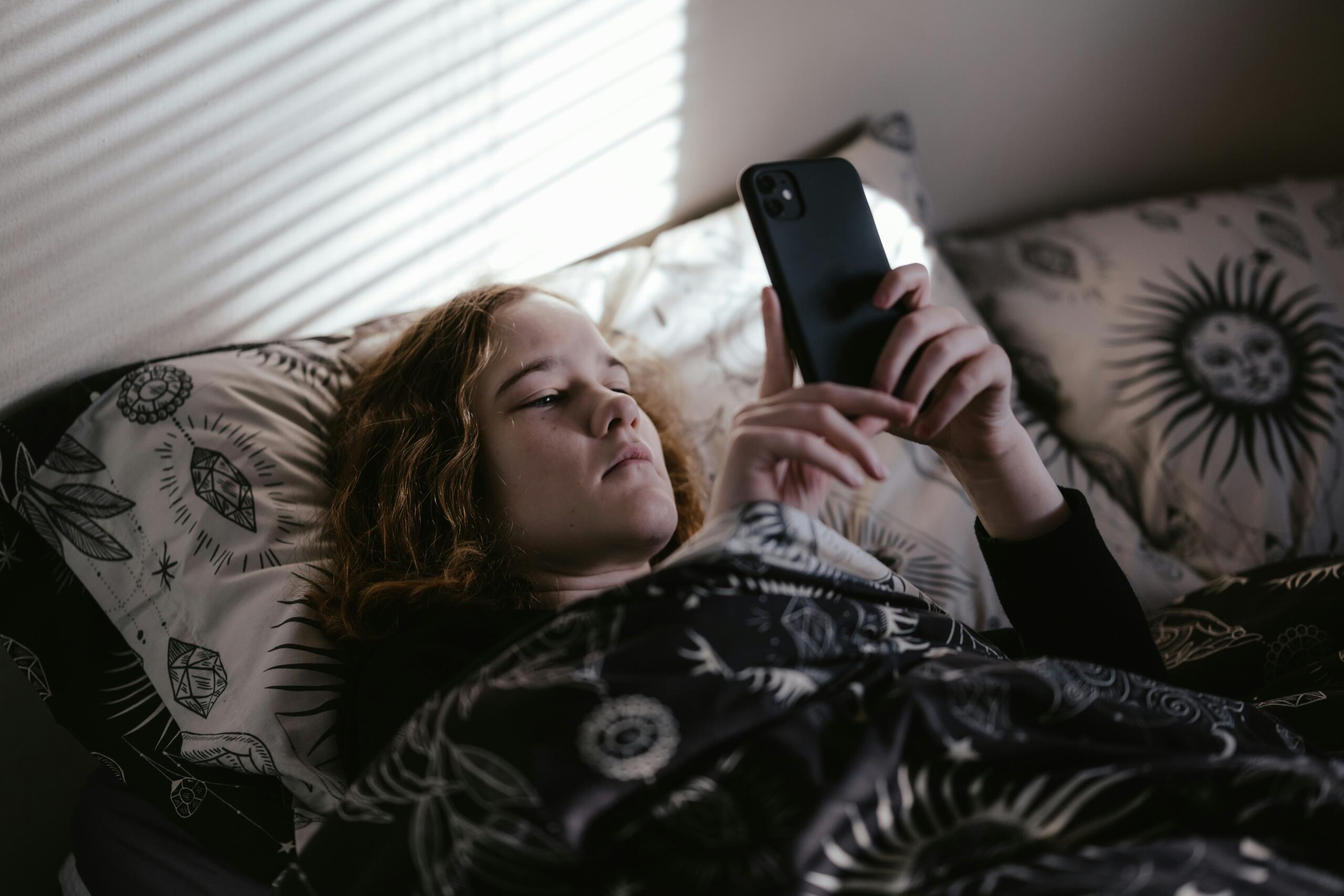 Παιδί χρησιμοποιεί smartphone ξαπλωμένο στο κρεβάτι κατά τις βραδινές ώρες, σε περιβάλλον υπνοδωματίου.