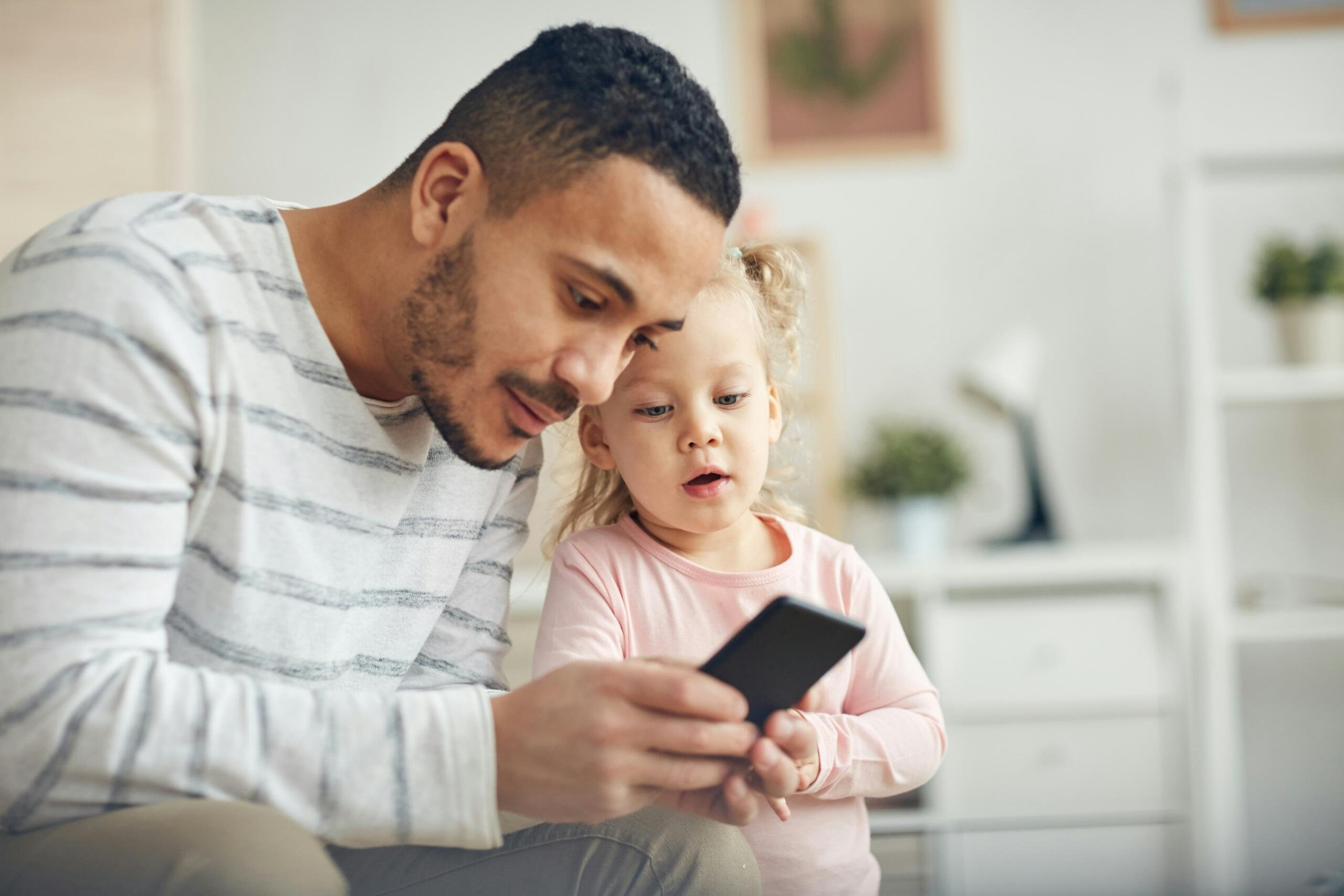 Πατέρας δείχνει περιεχόμενο σε smartphone σε μικρό παιδί, ενισχύοντας την ασφαλή και καθοδηγούμενη χρήση της τεχνολογίας στο σπίτι.