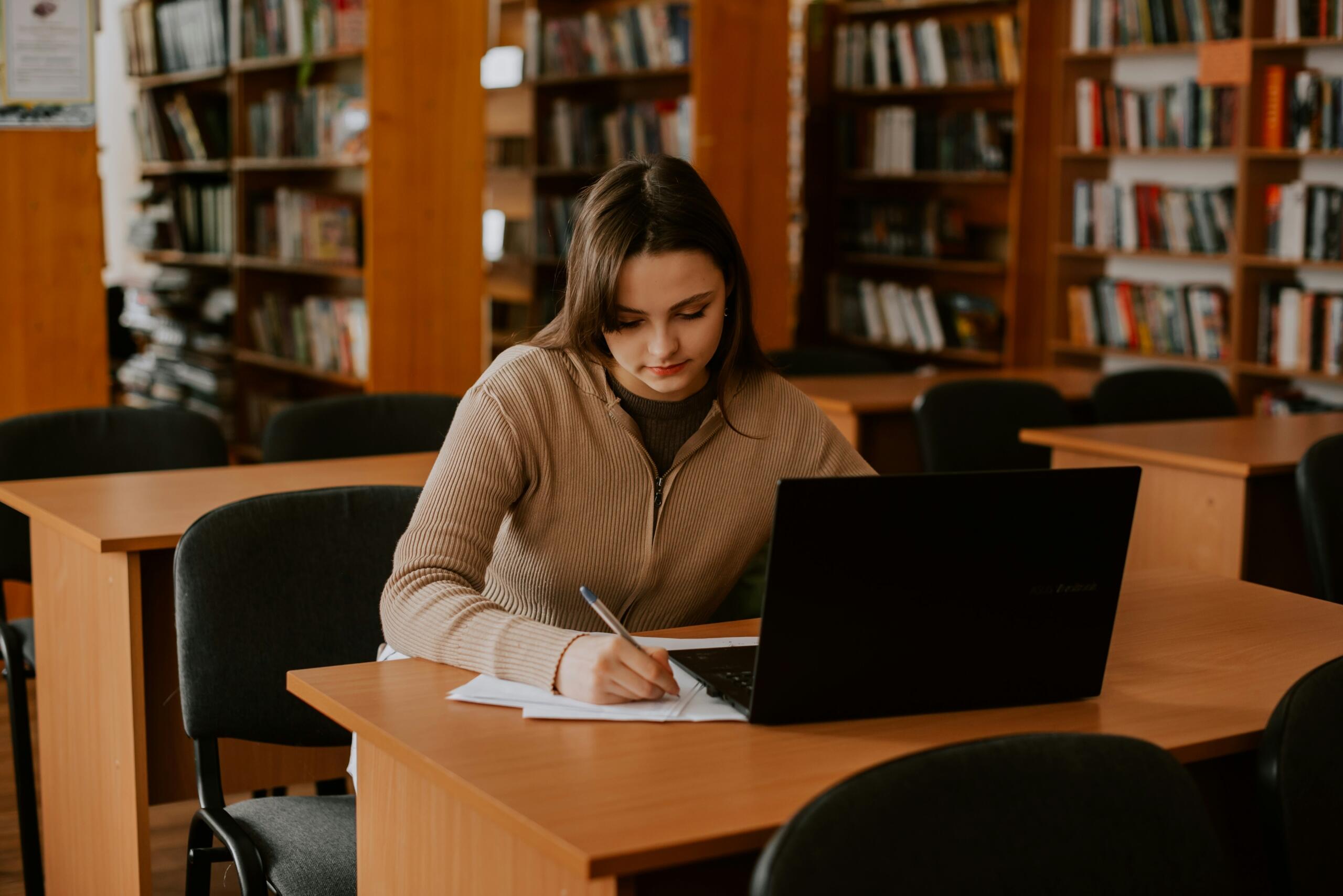 Φοιτήτρια γράφει σημειώσεις μπροστά από laptop σε βιβλιοθήκη με ξύλινα ράφια γεμάτα βιβλία