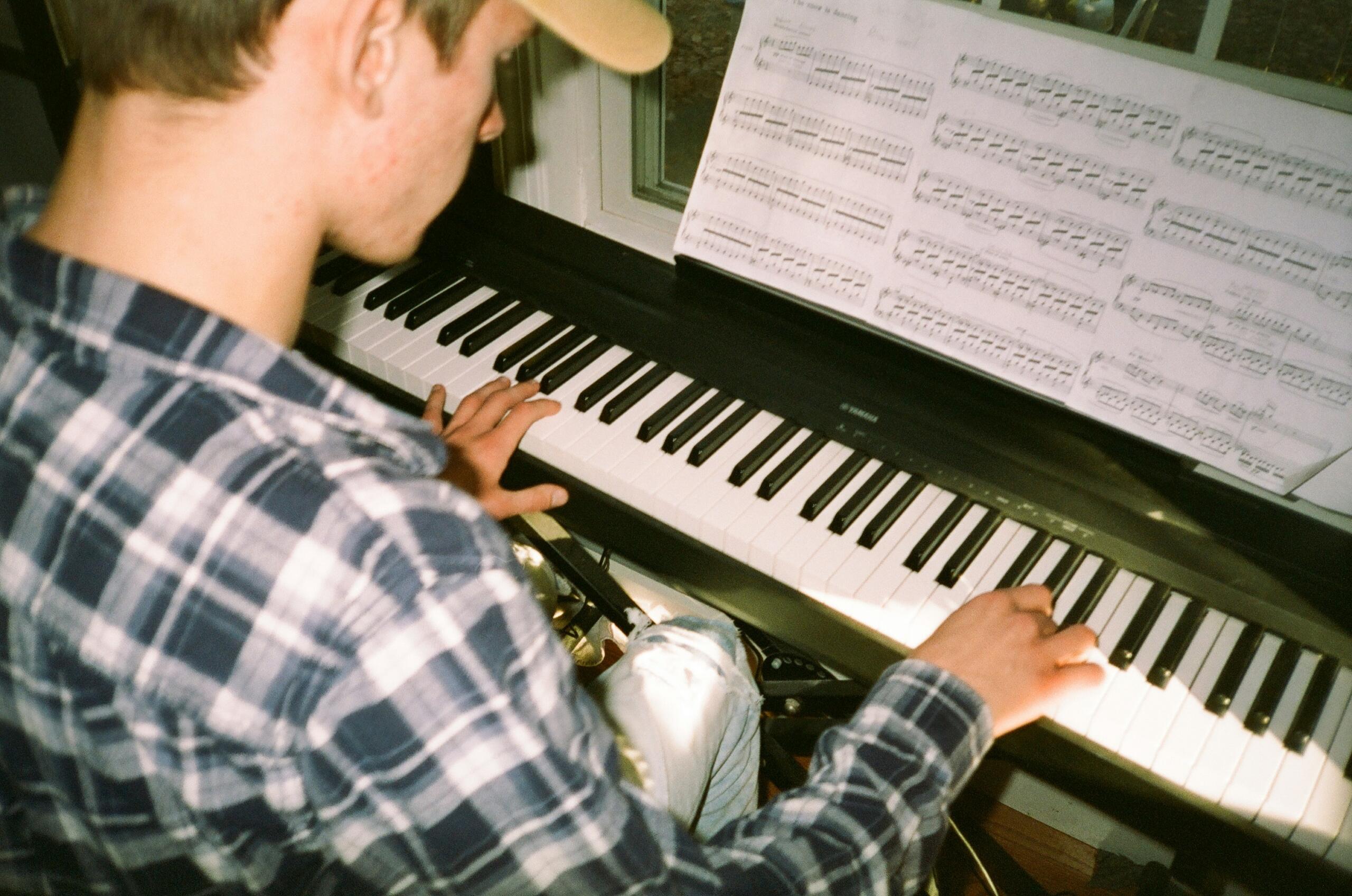 Νεαρός με καπέλο παίζει πιάνο Yamaha διαβάζοντας παρτιτούρα τοποθετημένη στη βάση, φορώντας καρό πουκάμισο σε μπλε και λευκές αποχρώσεις, με τα χέρια του πάνω στα πλήκτρα δίπλα σε παράθυρο με φυσικό φως
