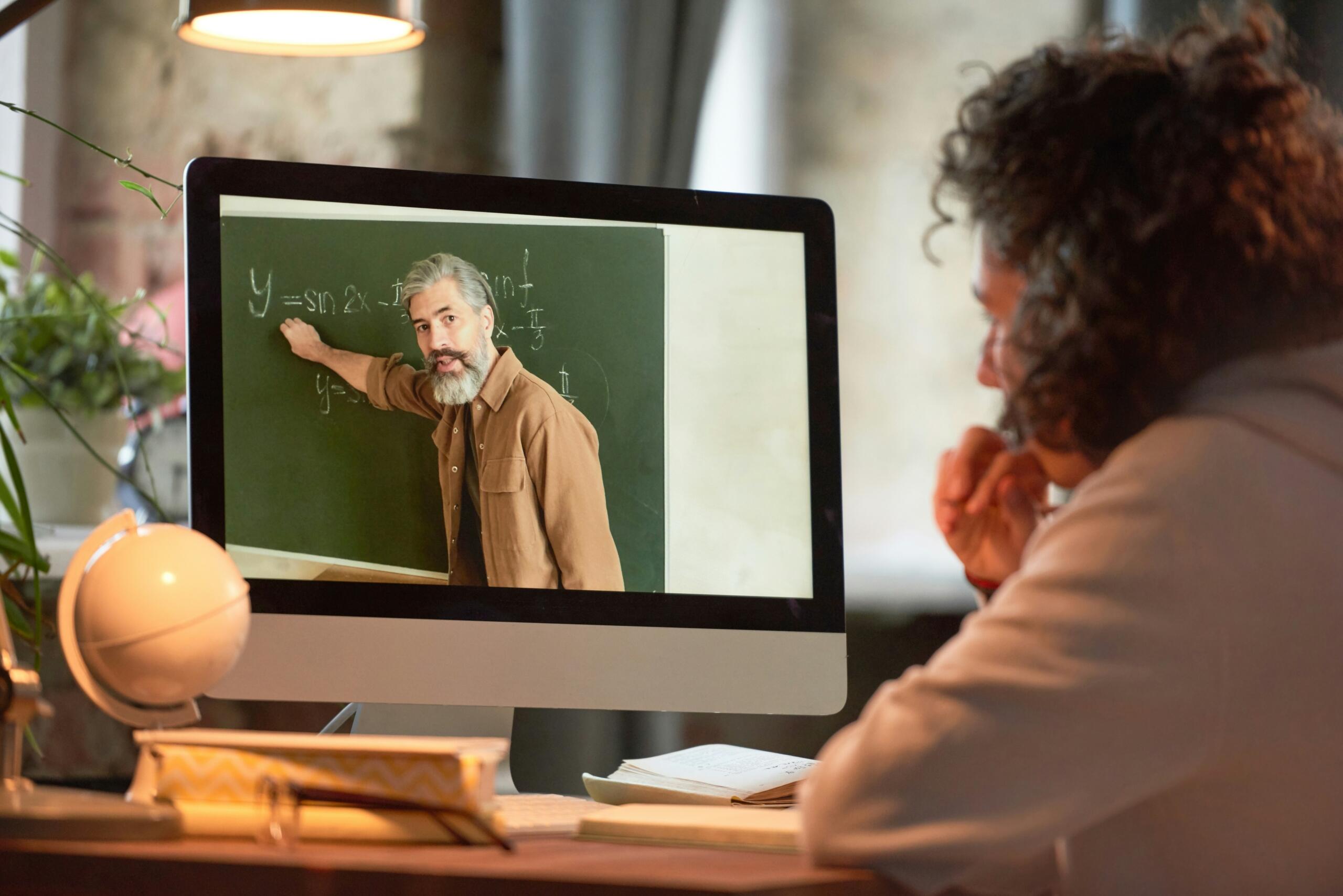Μαθητής παρακολουθεί online μάθημα μαθηματικών σε οθόνη υπολογιστή, με τον καθηγητή να εξηγεί τριγωνομετρικές συναρτήσεις σε μαυροπίνακα