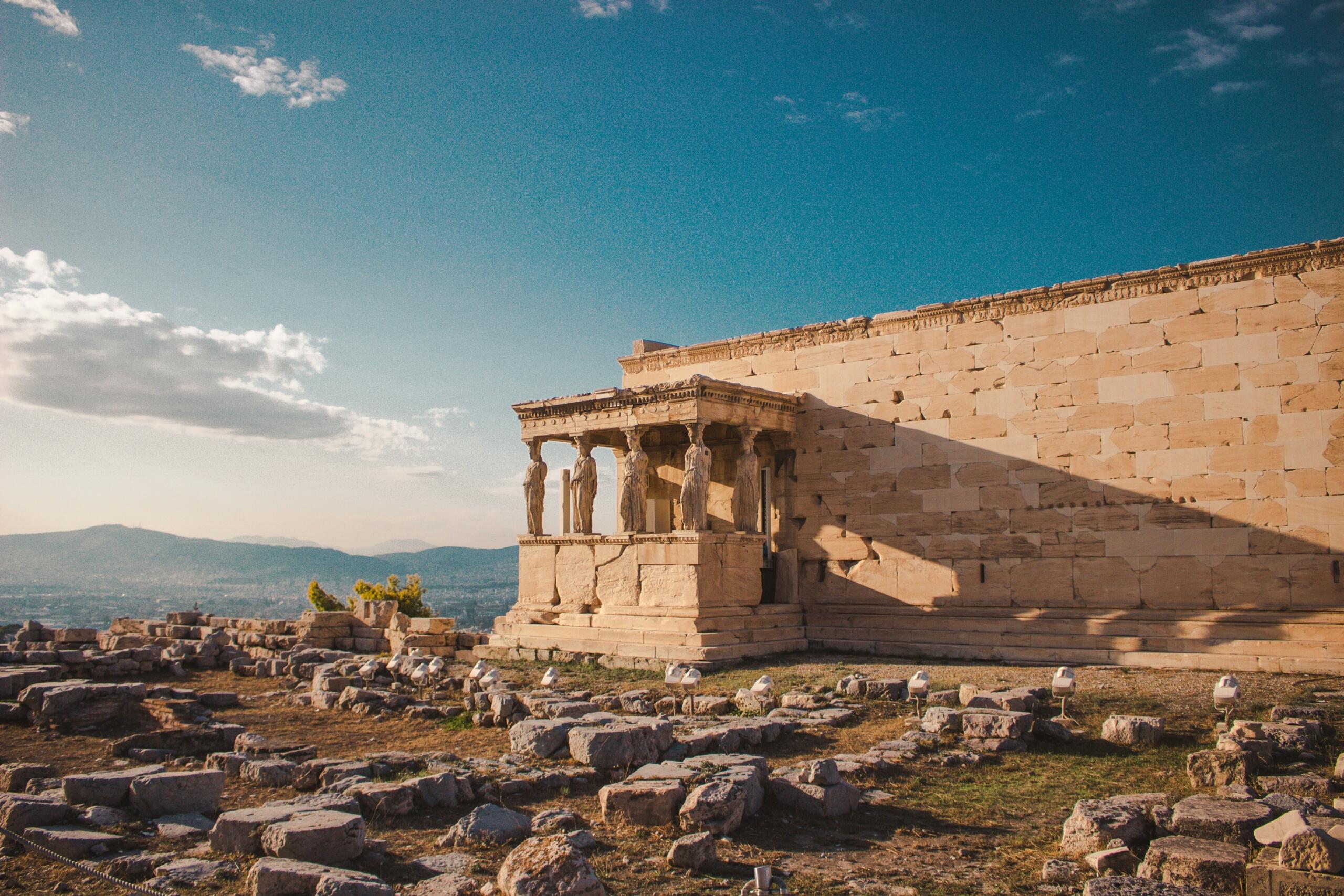 Το Ερέχθειο στην Ακρόπολη με τις έξι Καρυάτιδες στο πρόπυλο, φωτισμένο από το ηλιοβασίλεμα, με πέτρινο τείχος και σκιά να απλώνεται δίπλα, θέα σε βουνά στο βάθος