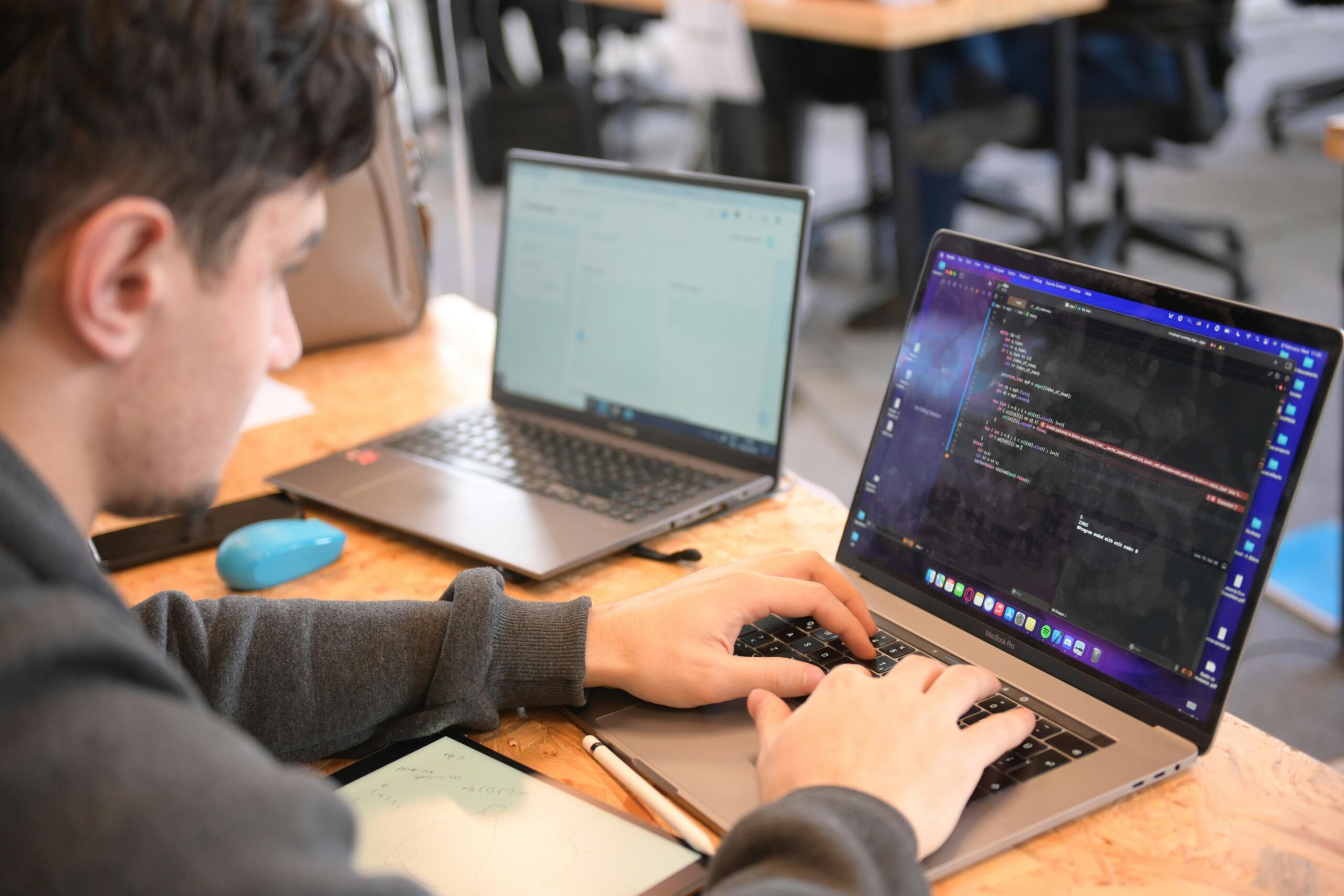 Άντρας πληκτρολογεί σε MacBook Pro με κώδικα στην οθόνη, δίπλα σε δεύτερο laptop σε ξύλινο γραφείο.