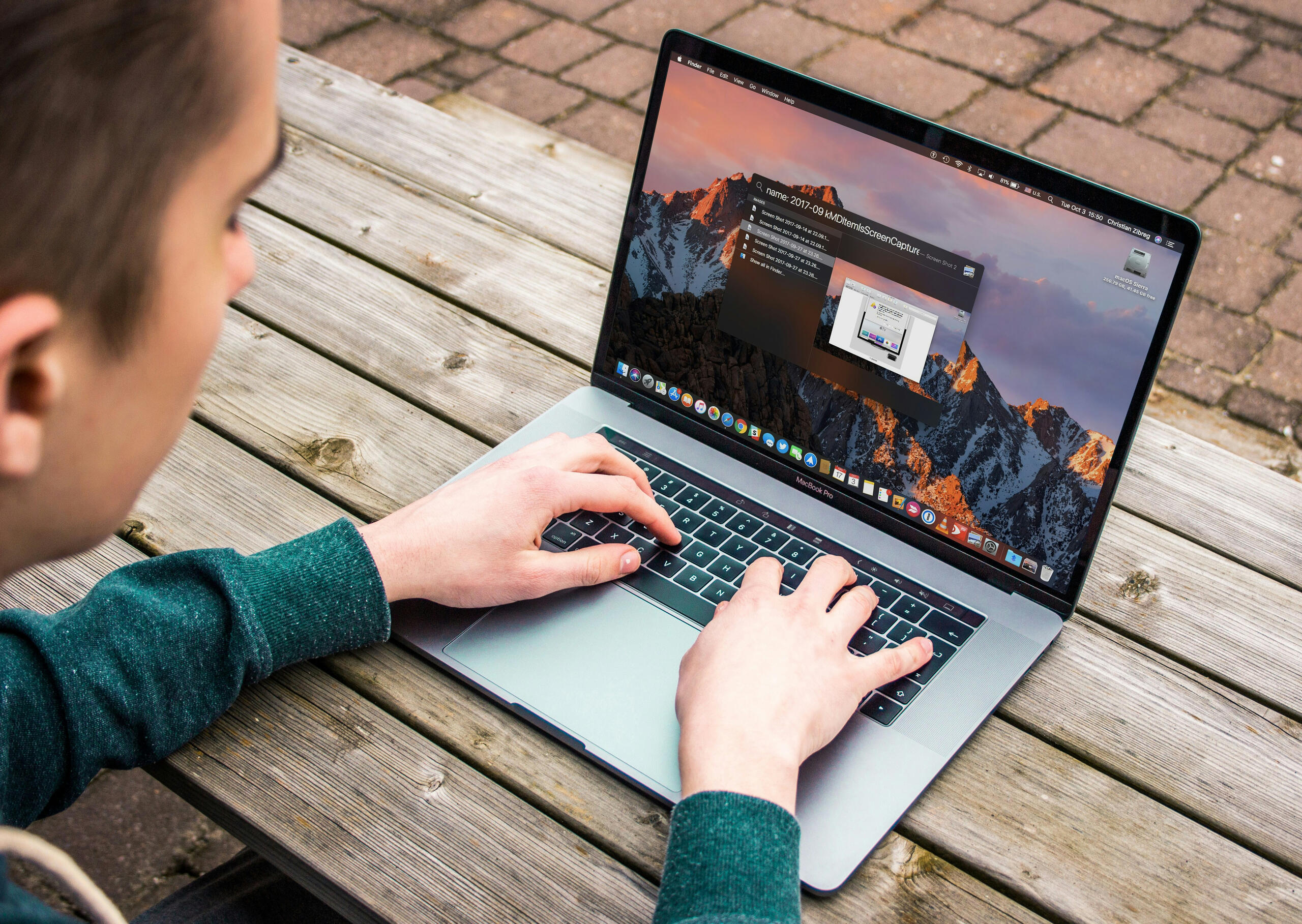 Κοντινό πλάνο χεριών που πληκτρολογούν σε MacBook Pro με ανοιχτή αναζήτηση Spotlight πάνω σε ξύλινο τραπέζι εξωτερικού χώρου.
