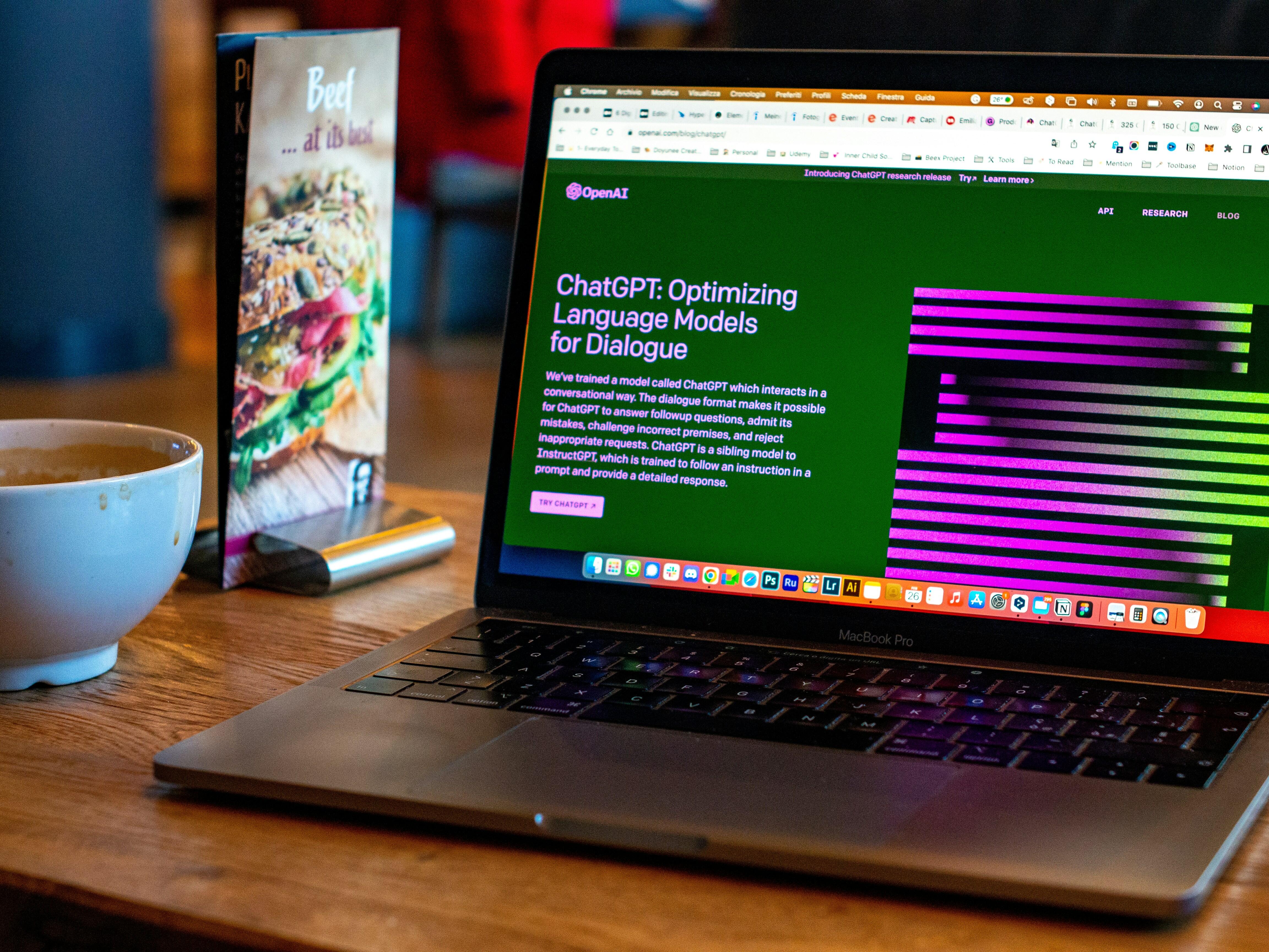 MacBook Pro σε ξύλινο τραπέζι με ανοιχτή ιστοσελίδα που παρουσιάζει το ChatGPT, δίπλα σε φλιτζάνι καφέ.