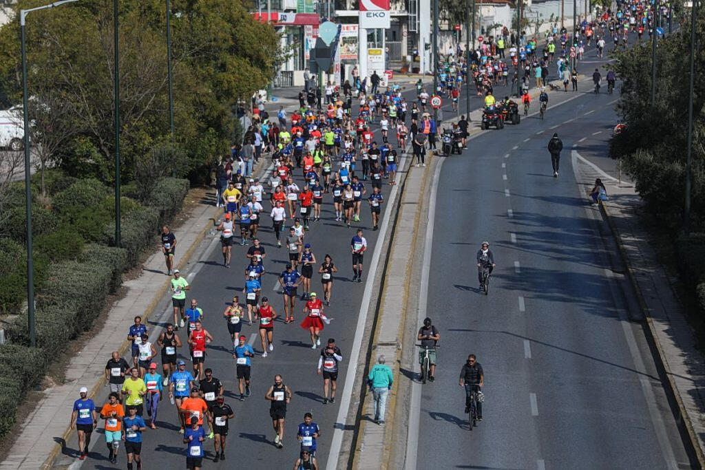 Αεροφωτογραφία δρομέων που τρέχουν σε φαρδύ δρόμο πόλης, με ποδηλάτες να παρακολουθούν τον αγώνα δίπλα τους.