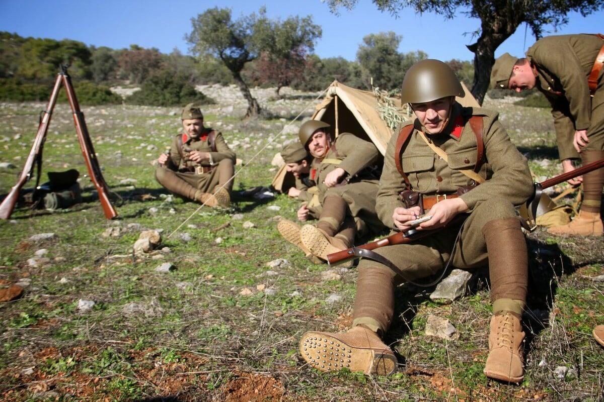 Άνδρες με αναπαράσταση στολών του ελληνικού στρατού της εποχής 1940-41, κάθονται σε εξωτερικό χώρο με όπλα και σκηνή εκστρατείας στο φόντο.