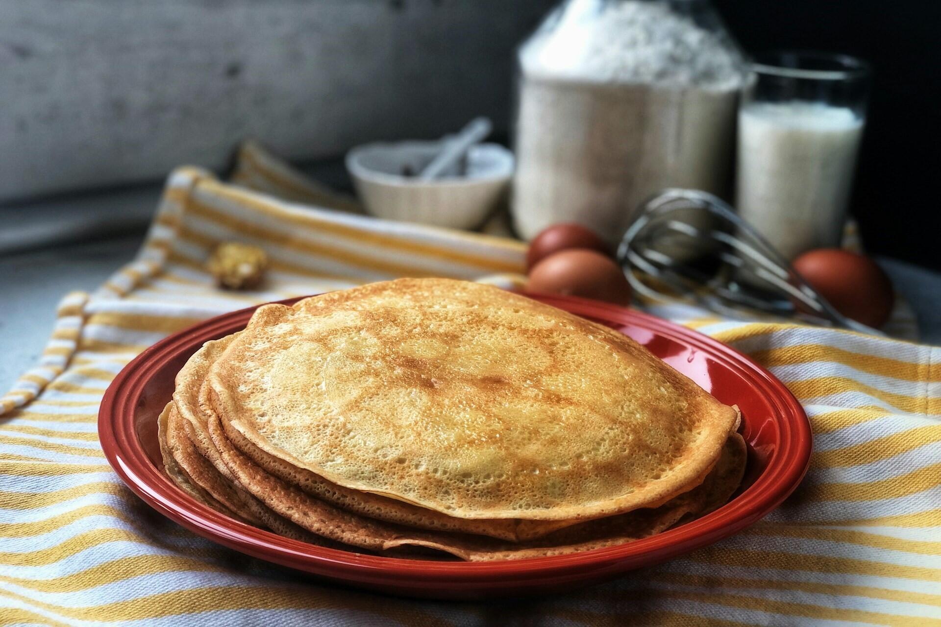 Stack of French crêpes on a red plate with flour and eggs in the background