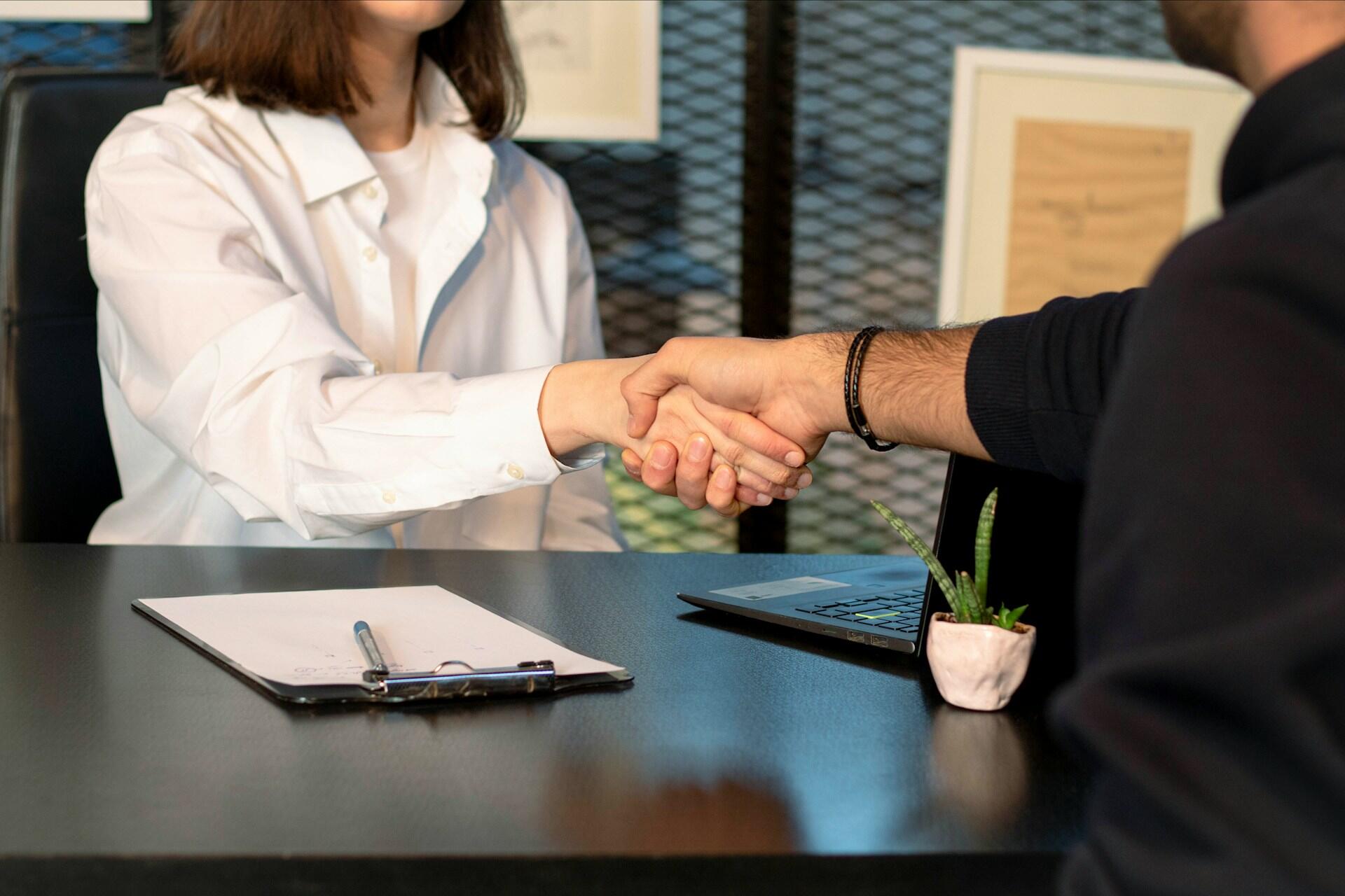 Two people shaking hands across an office desk with a laptop and clipboard