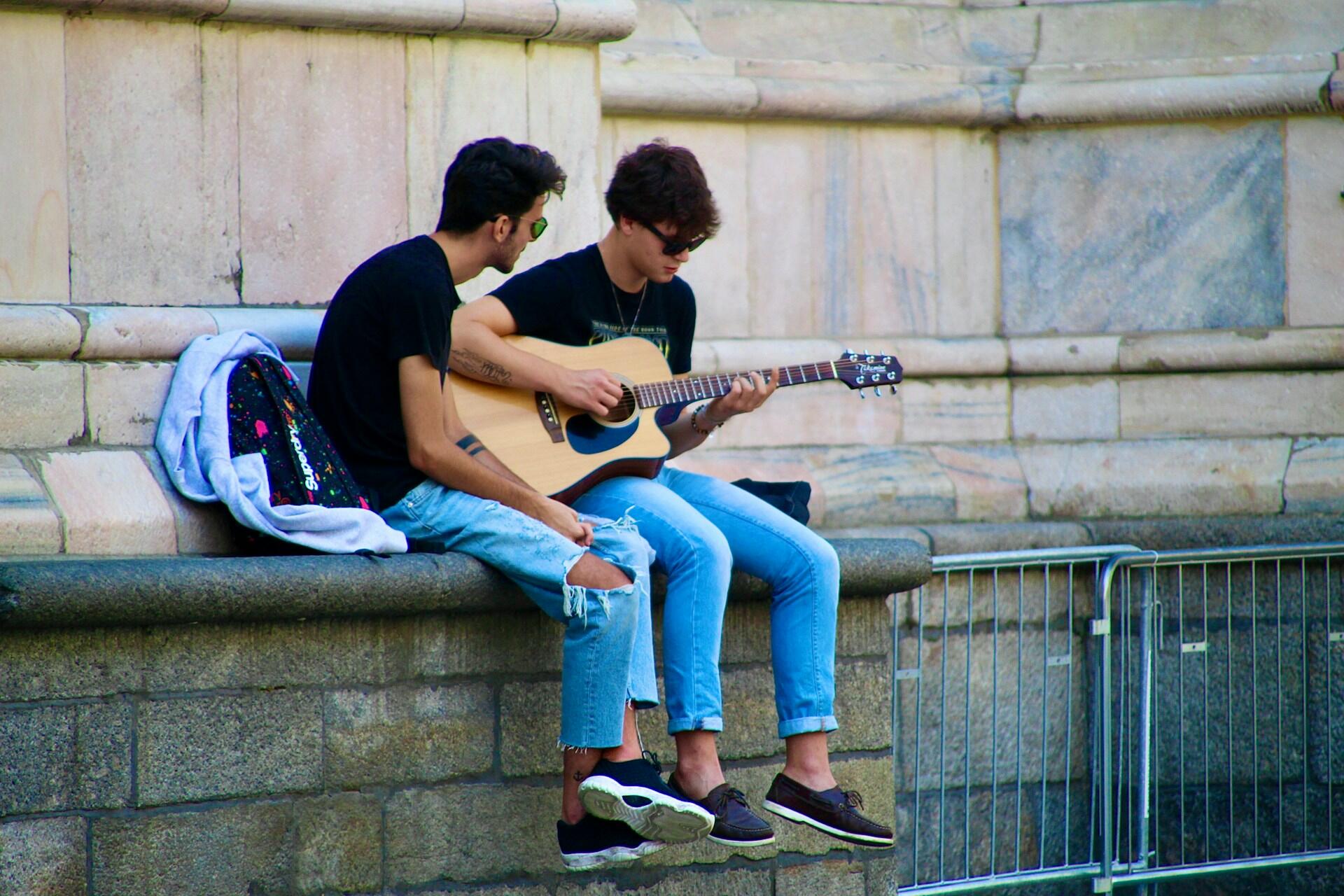 Two young men sitting on a stone ledge playing an acoustic guitar outdoors