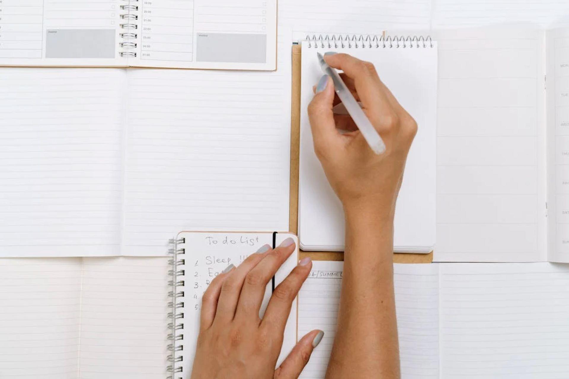 Person writing in notebook on desk with planners and to-do list.