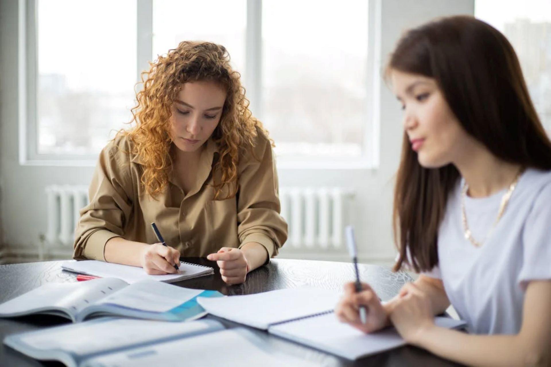 Two students writing in notebooks, studying together at a table with textbooks