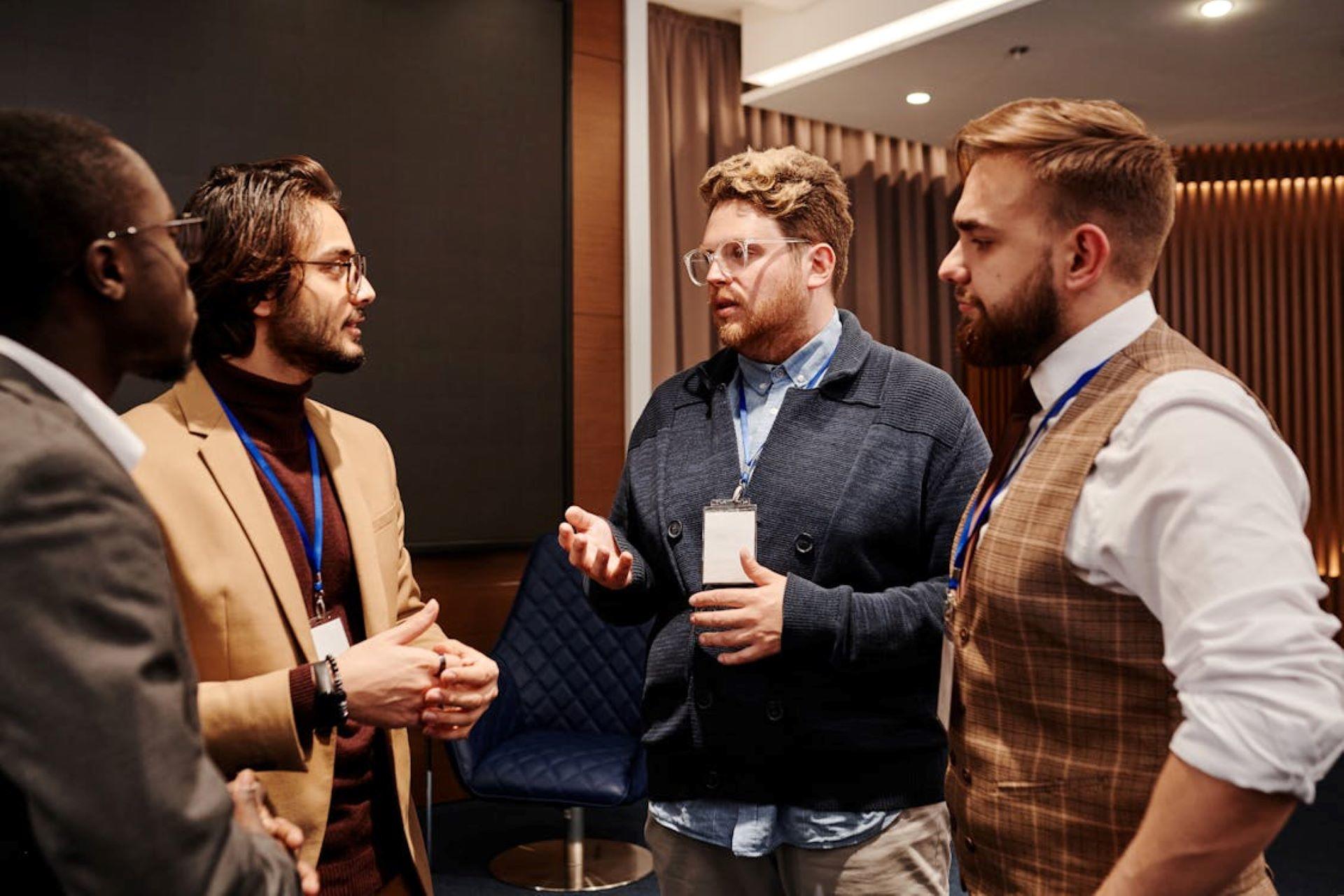 Four men wearing lanyards discuss something at a conference, practicing their speaking skills