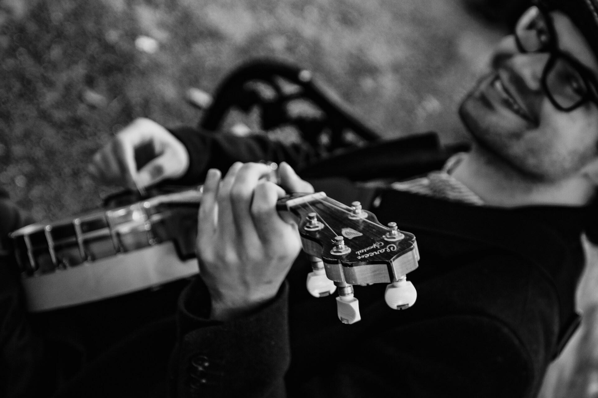 A smiling musician plays a banjo, partially in focus, with textures of a park setting blurred in black and white.