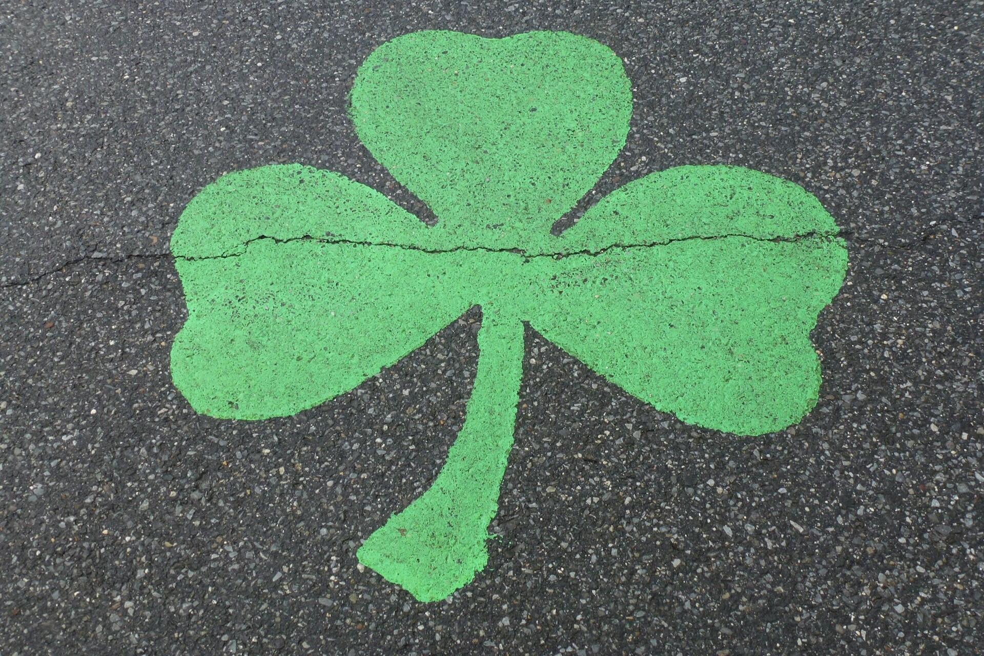 A large green shamrock painted on asphalt, featuring three heart-shaped leaves and a central stem, with a visible crack in the surface.