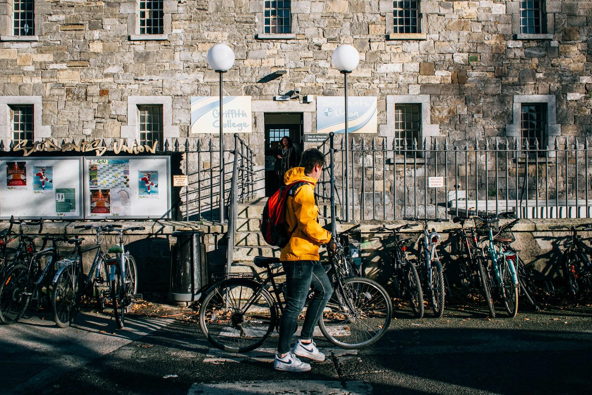 A man in a yellow coat outside a college with bicycles.