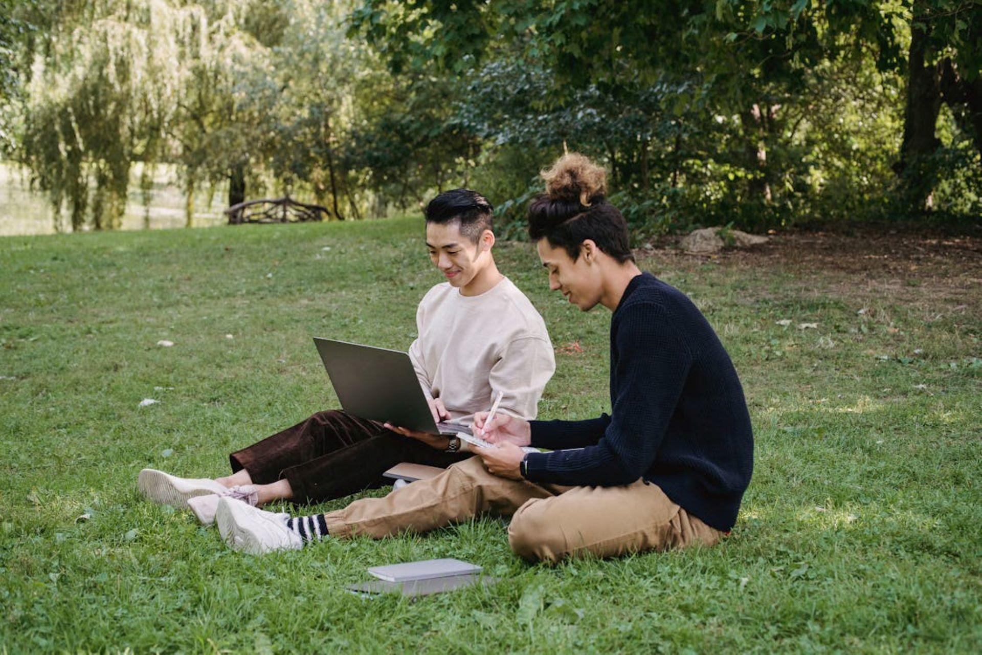 Two students studying in the park, one using a laptop and smiling, the other taking notes