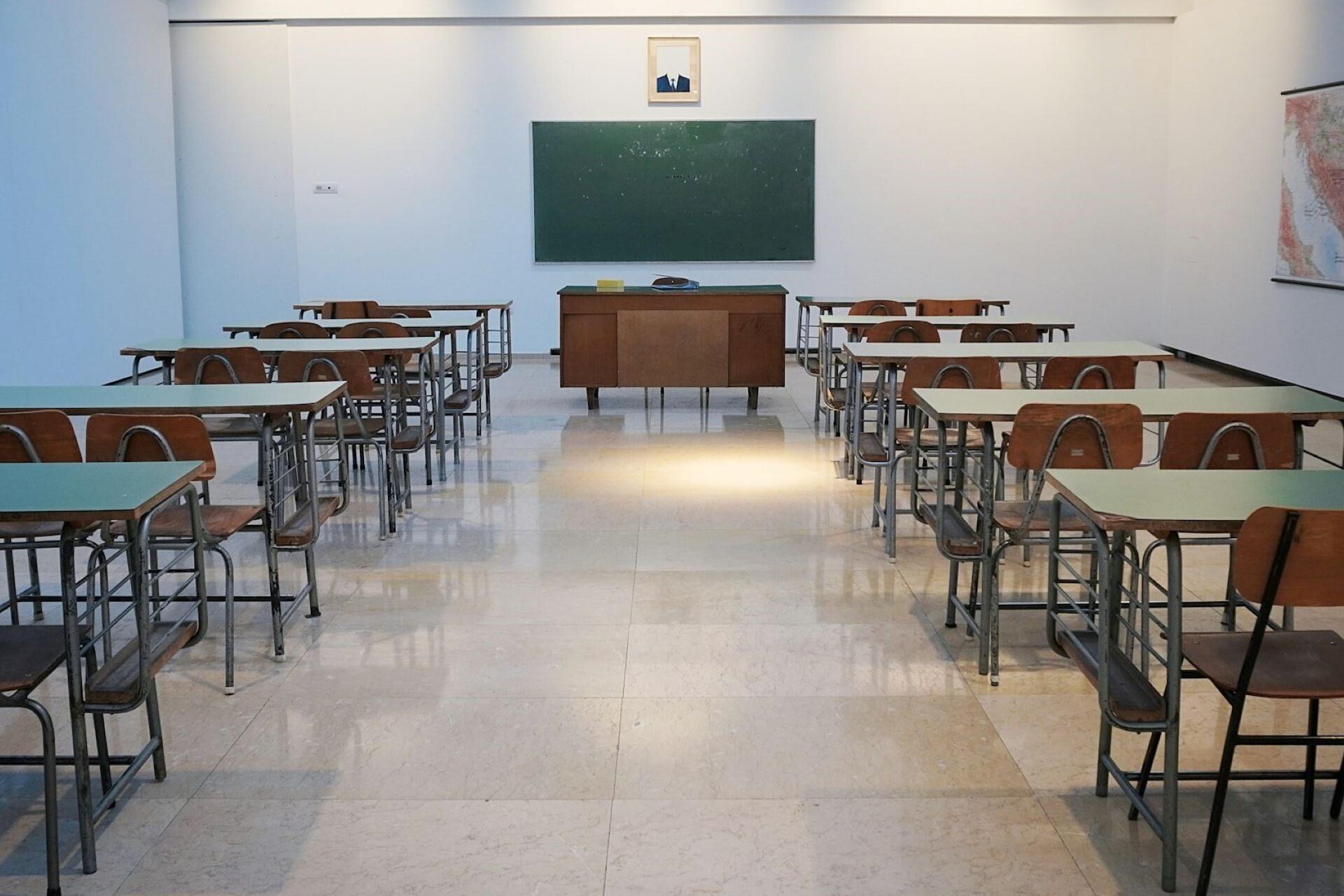 An empty classroom with wooden desks and chairs, a chalkboard, and a teacher's desk at the front. A portrait hangs above the chalkboard.