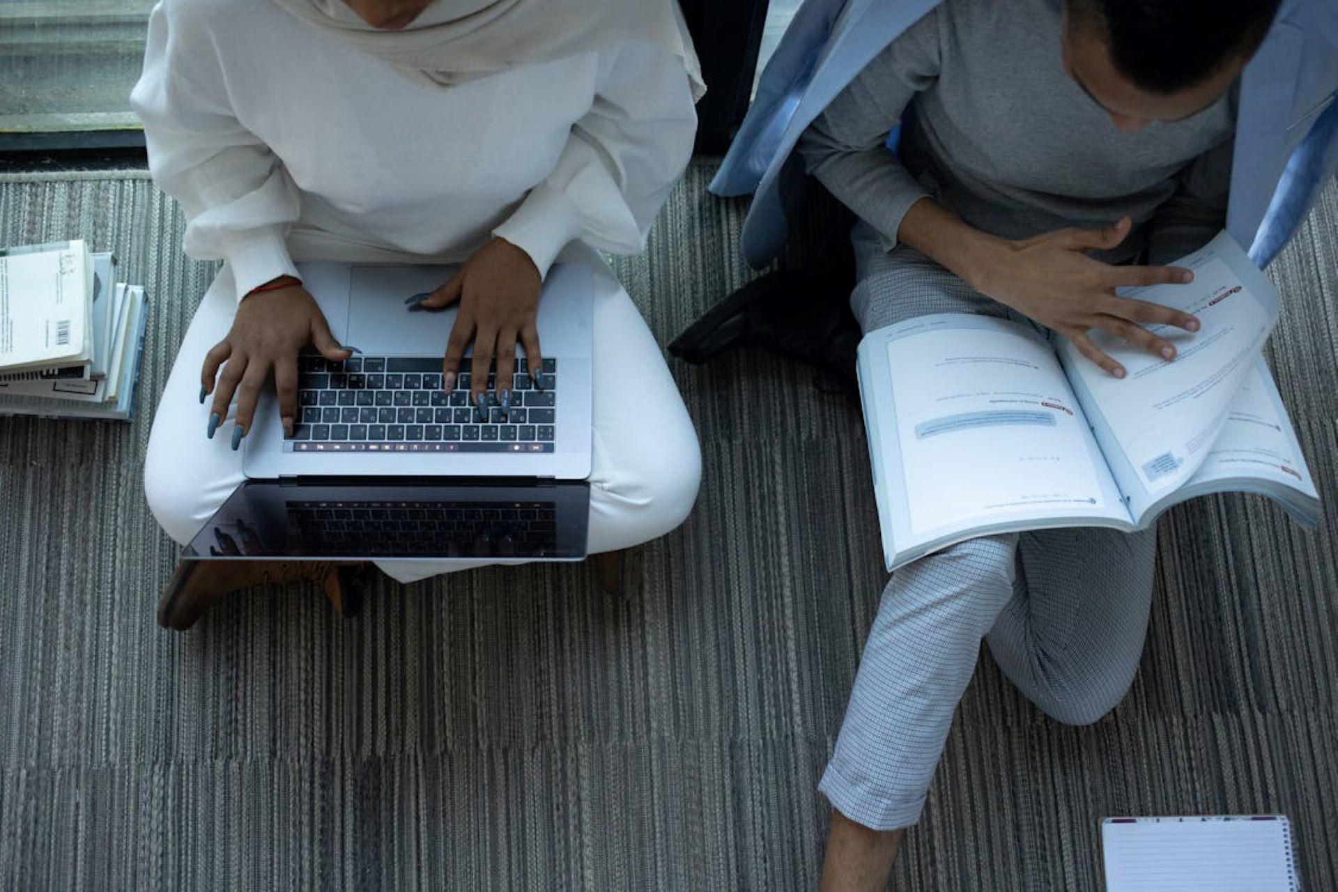 Two students studying together on the floor with a laptop and a book