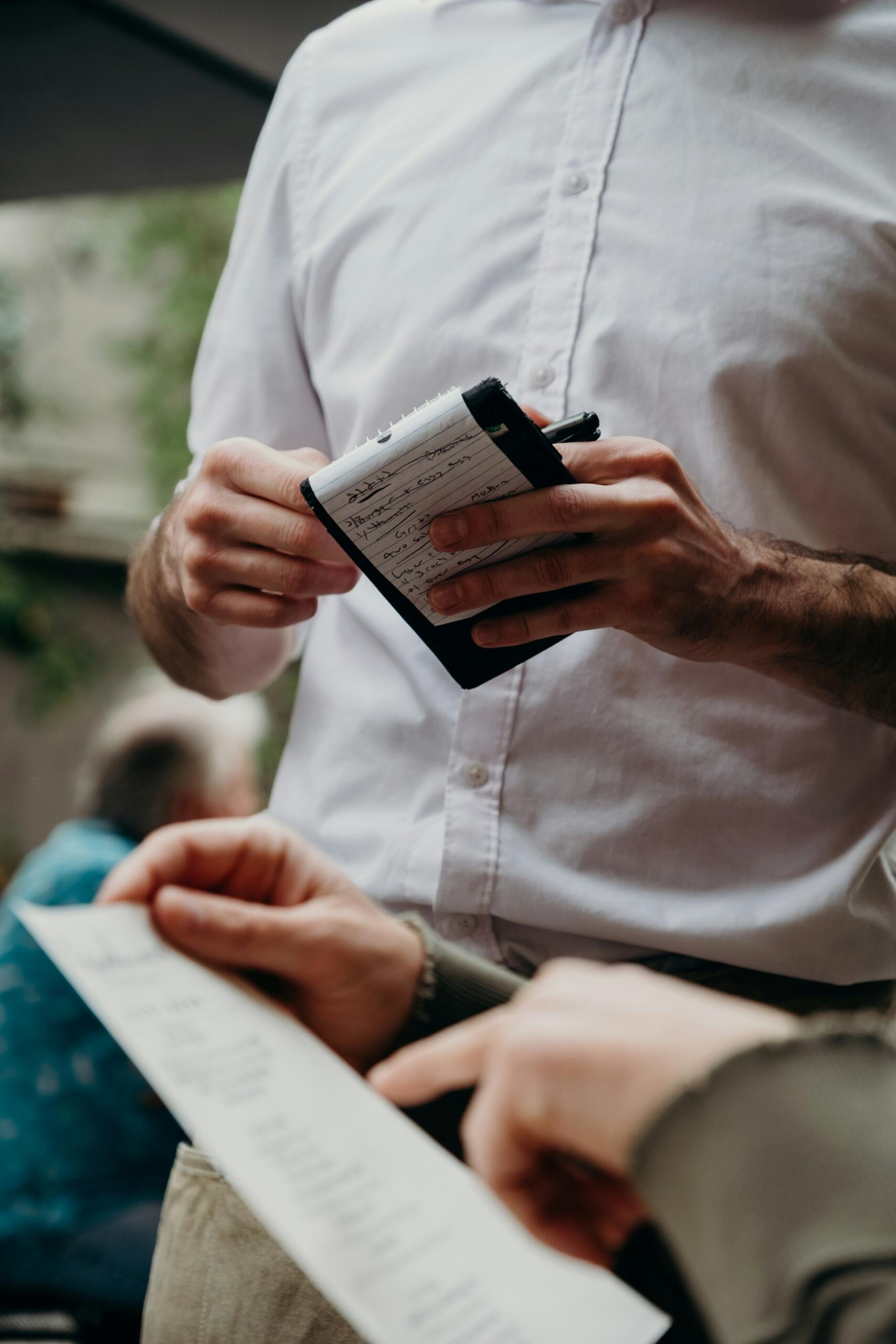 Person holding a small notepad and pen while another person points at a paper