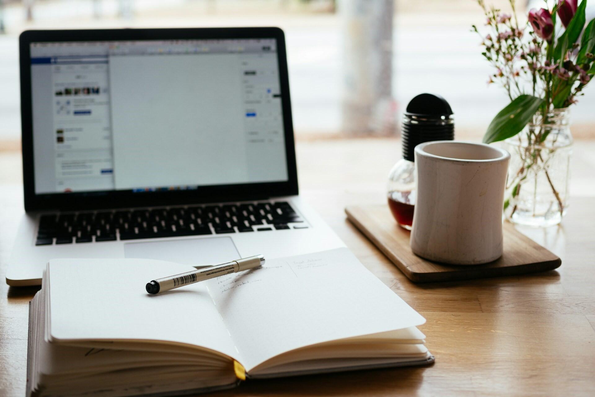 Open notebook and pen in front of a laptop, with a coffee cup and flowers on a wooden table.