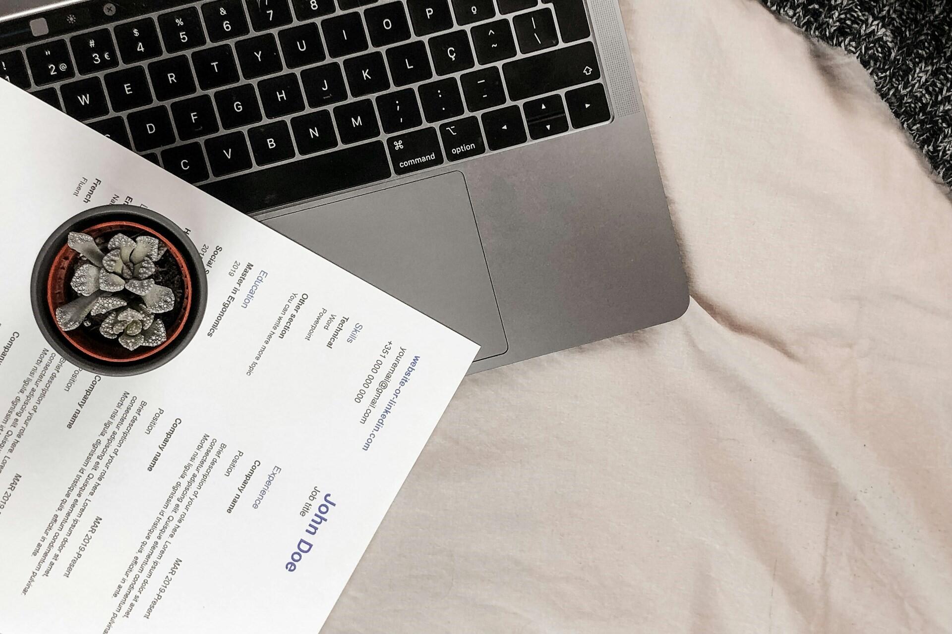 Laptop keyboard beside printed documents and a small potted plant on a desk.