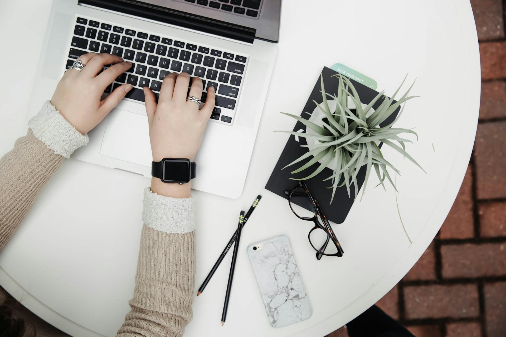 Hands typing on a laptop at a white table with pencils, glasses, a phone, and a plant nearby.