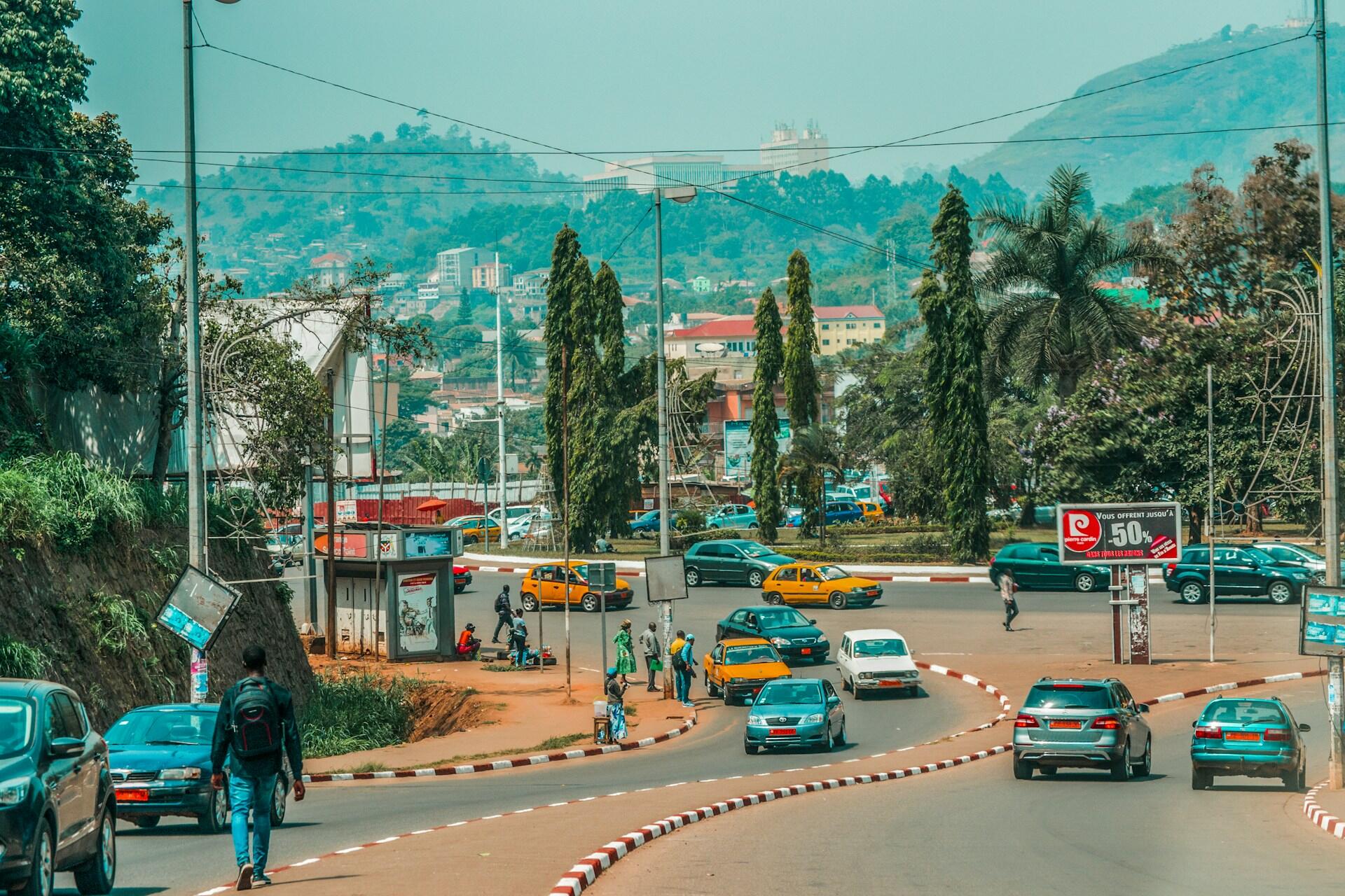 City road with cars, pedestrians, trees, hills, and buildings in the background