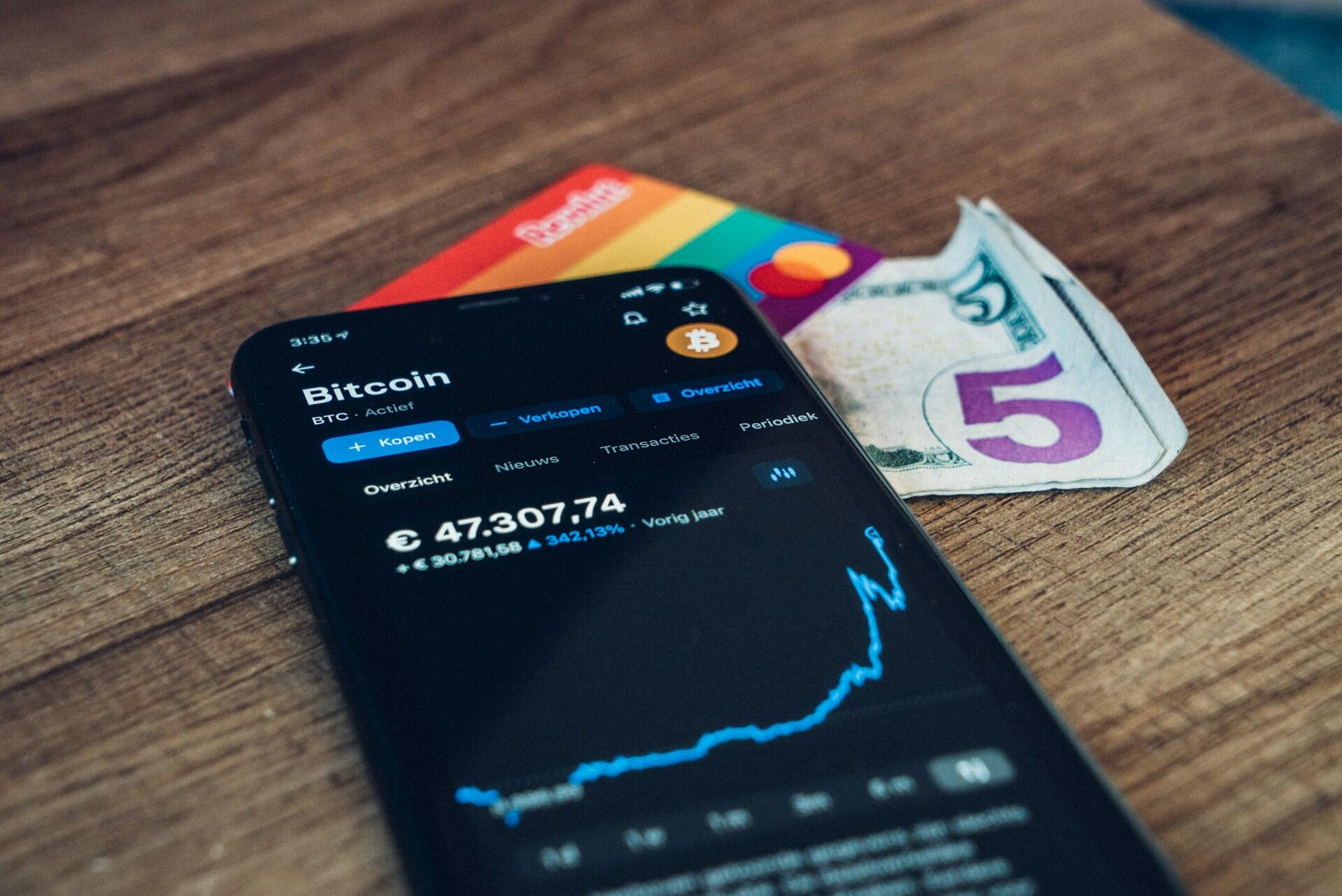 Smartphone displaying a Bitcoin price chart beside cards and cash