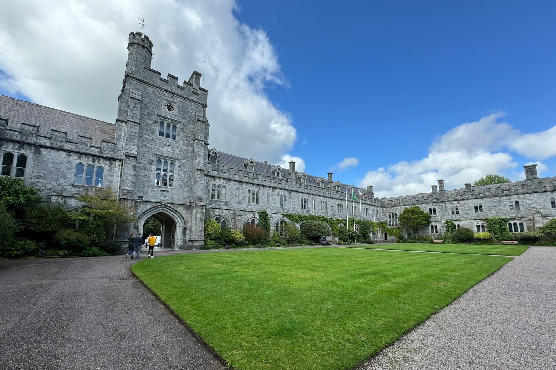 stone university building with courtyard lawn and people walking near entrance
