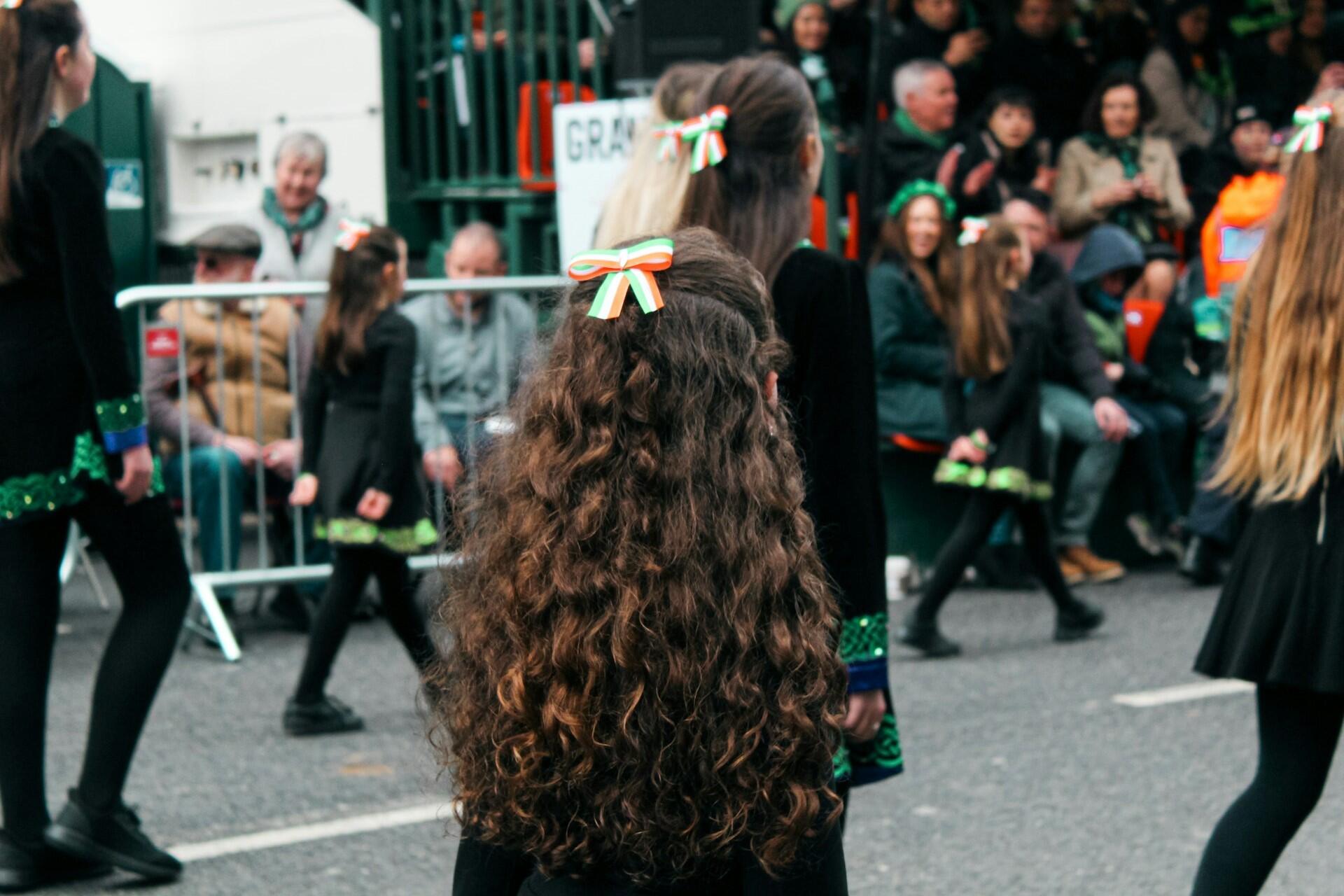 Young girls dancing in a street parade with Irish tricolour ribbons in their hair while a crowd watches