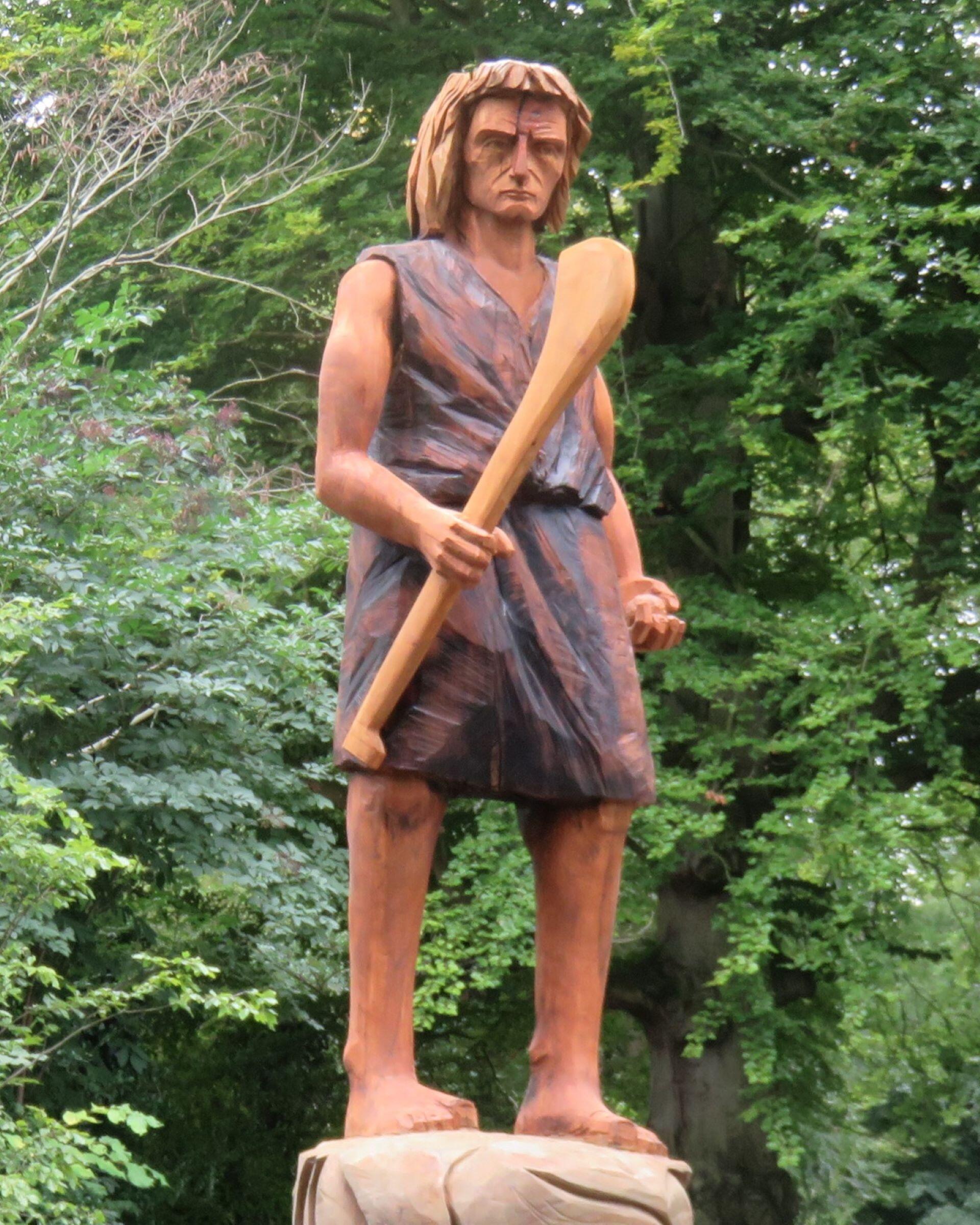 Wooden statue of a man holding a stick standing outdoors with trees in the background