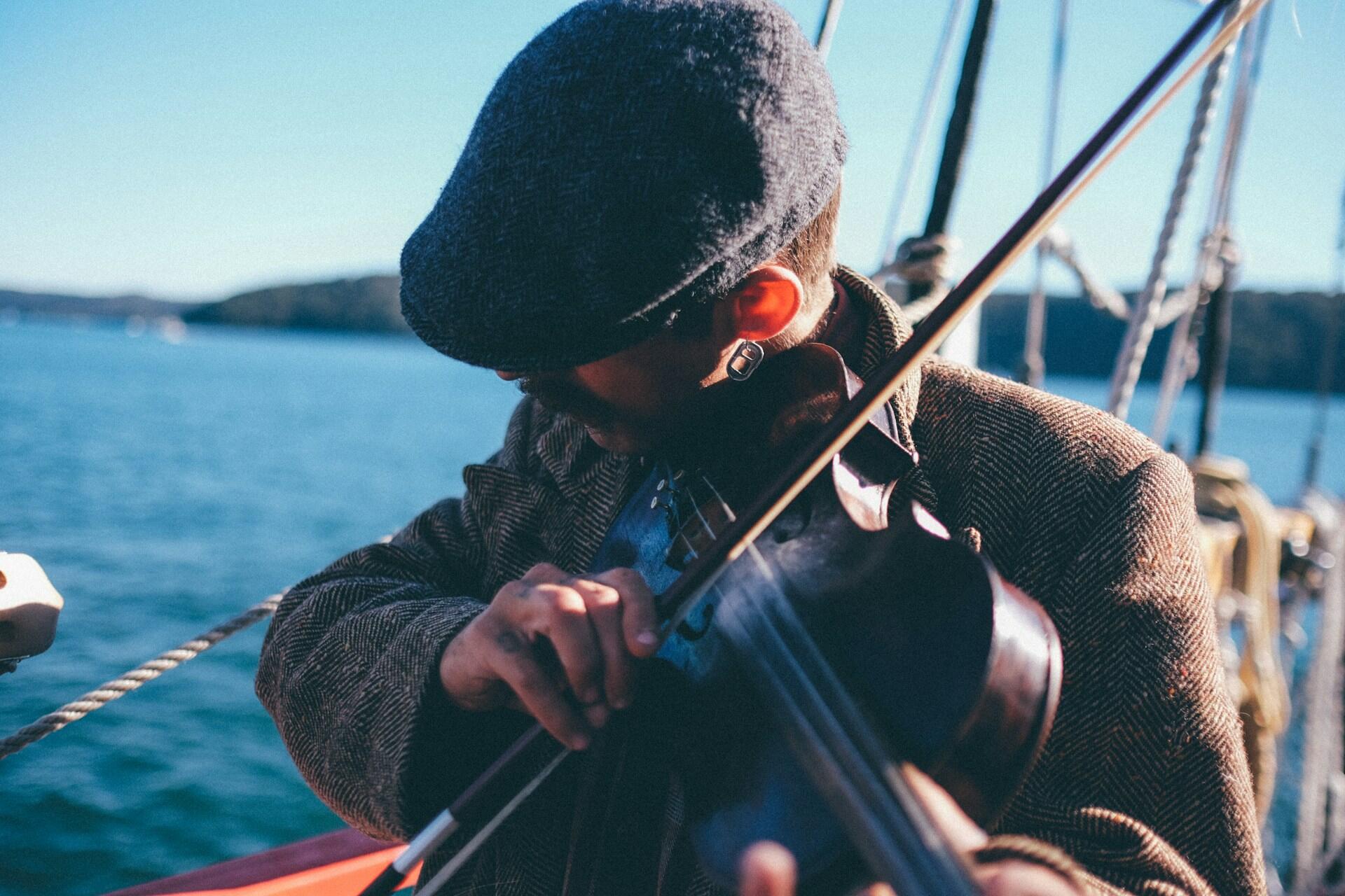 A person in a tweed jacket and flat cap plays the violin aboard a boat with a scenic water backdrop under a clear blue sky.