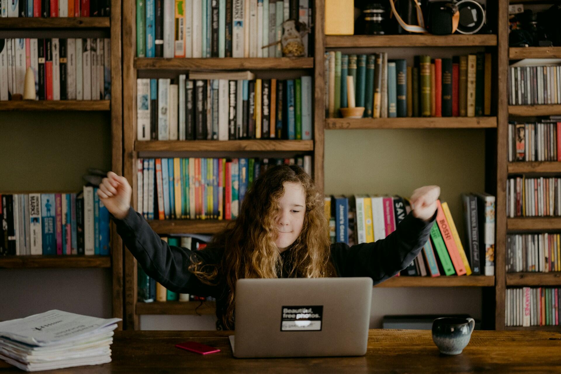 child celebrating while using a laptop in front of a bookshelf