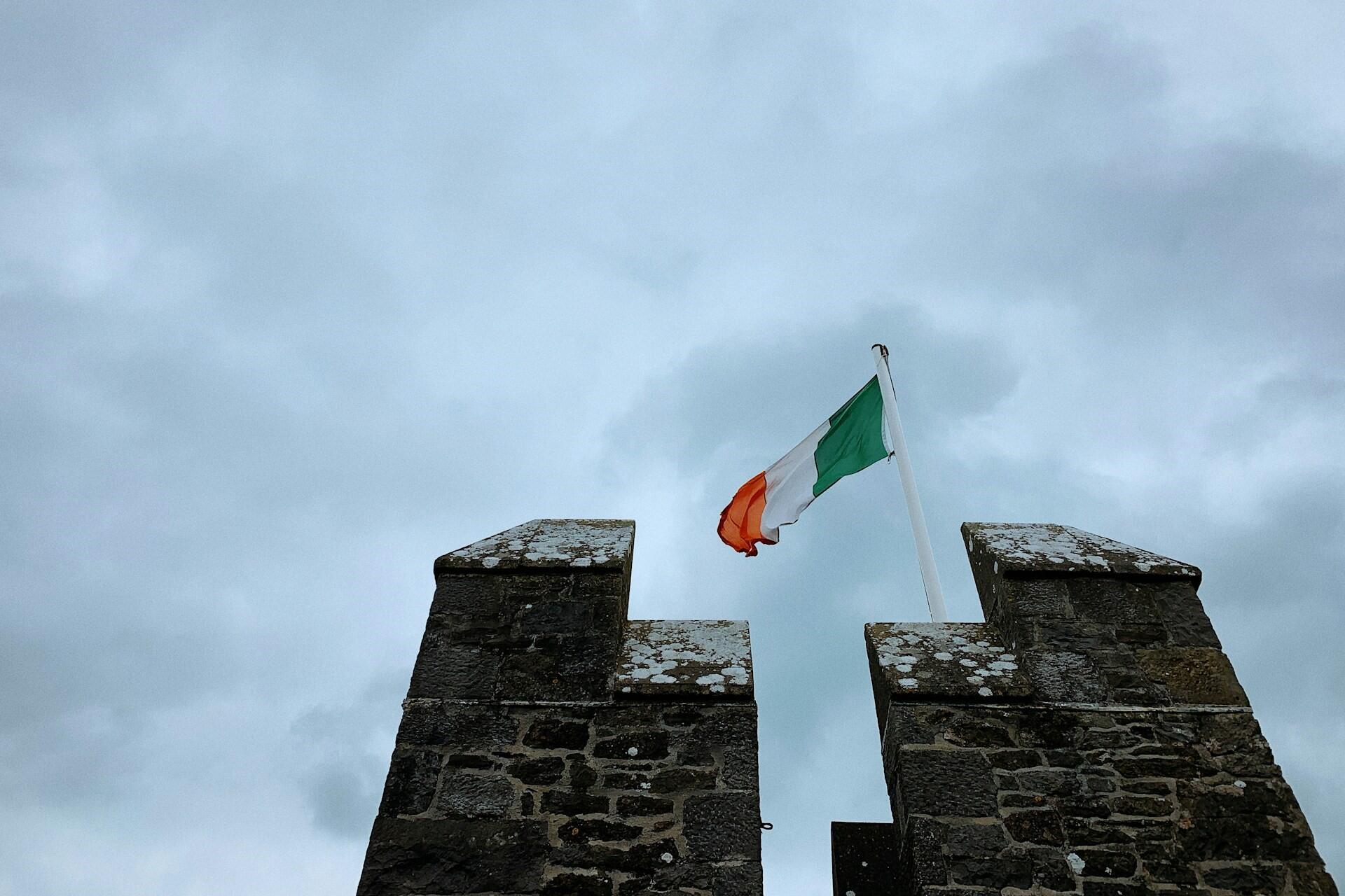 Irish flag flying above a stone castle wall under a cloudy sky