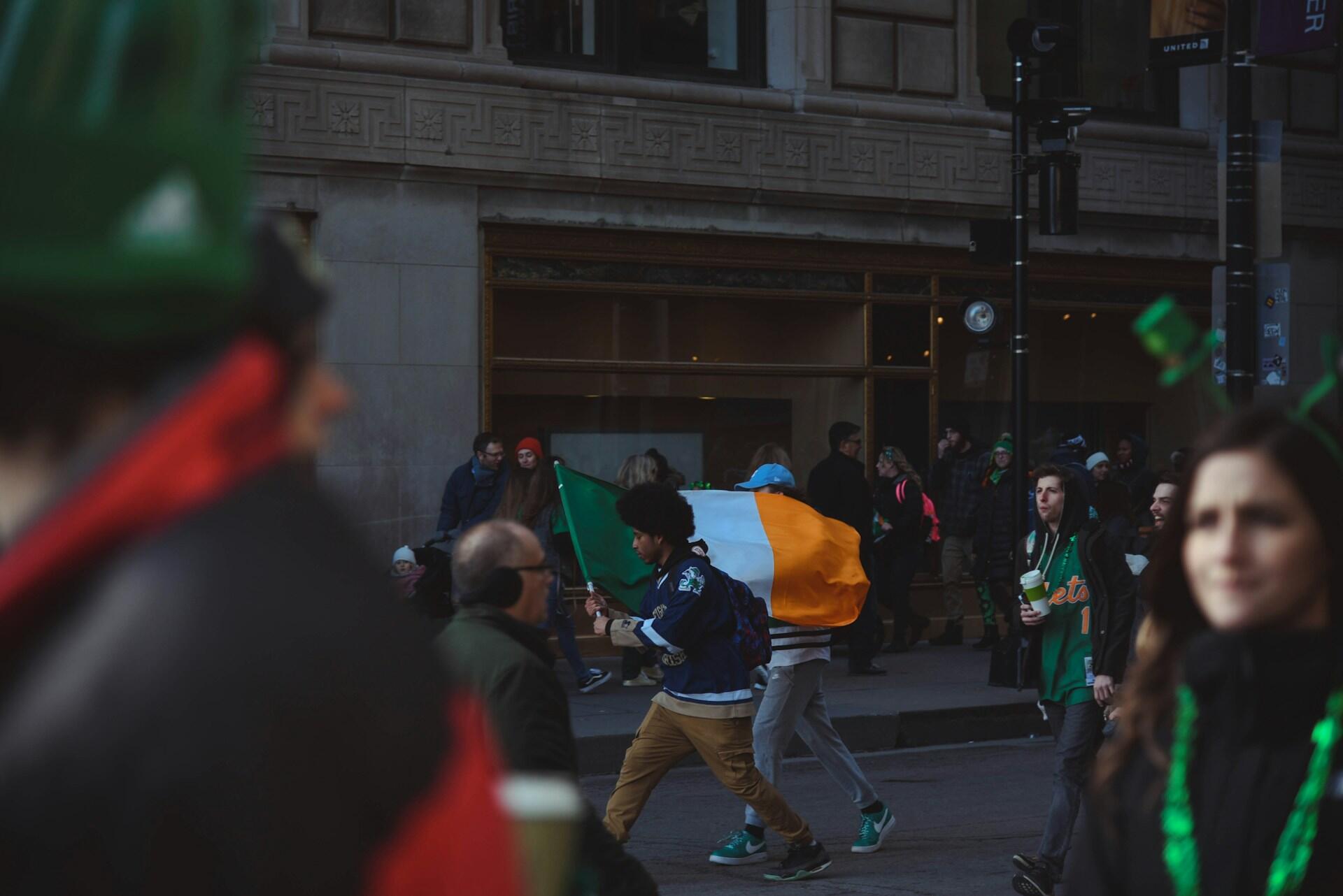 Crowd of people at a parade with an Irish flag in the foreground and buildings behind