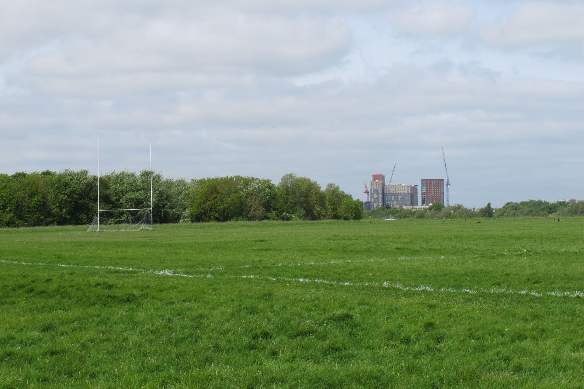 Empty grass GAA pitch with white goalposts, boundary markings and buildings in the distance