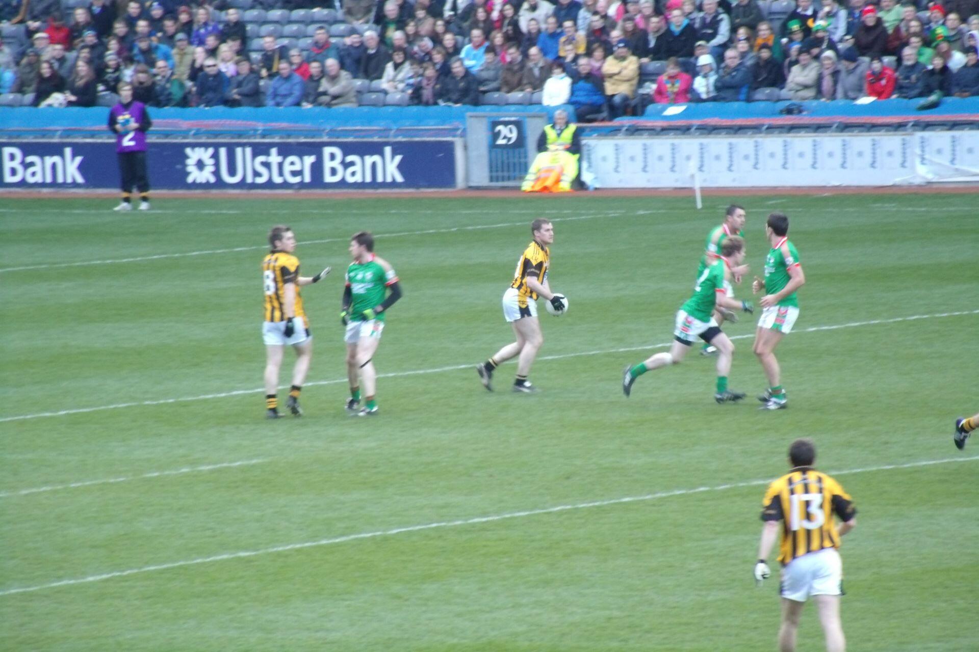 Gaelic football players in green, red, black and yellow kits during a match in a crowded stadium