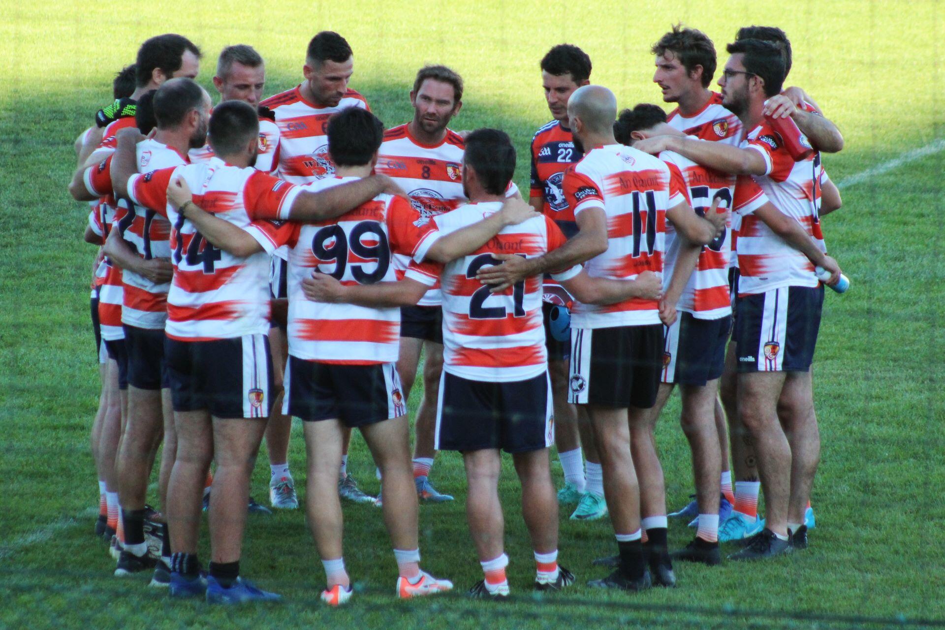 Gaelic football players in red-and-white jerseys standing in a team huddle on a grass pitch