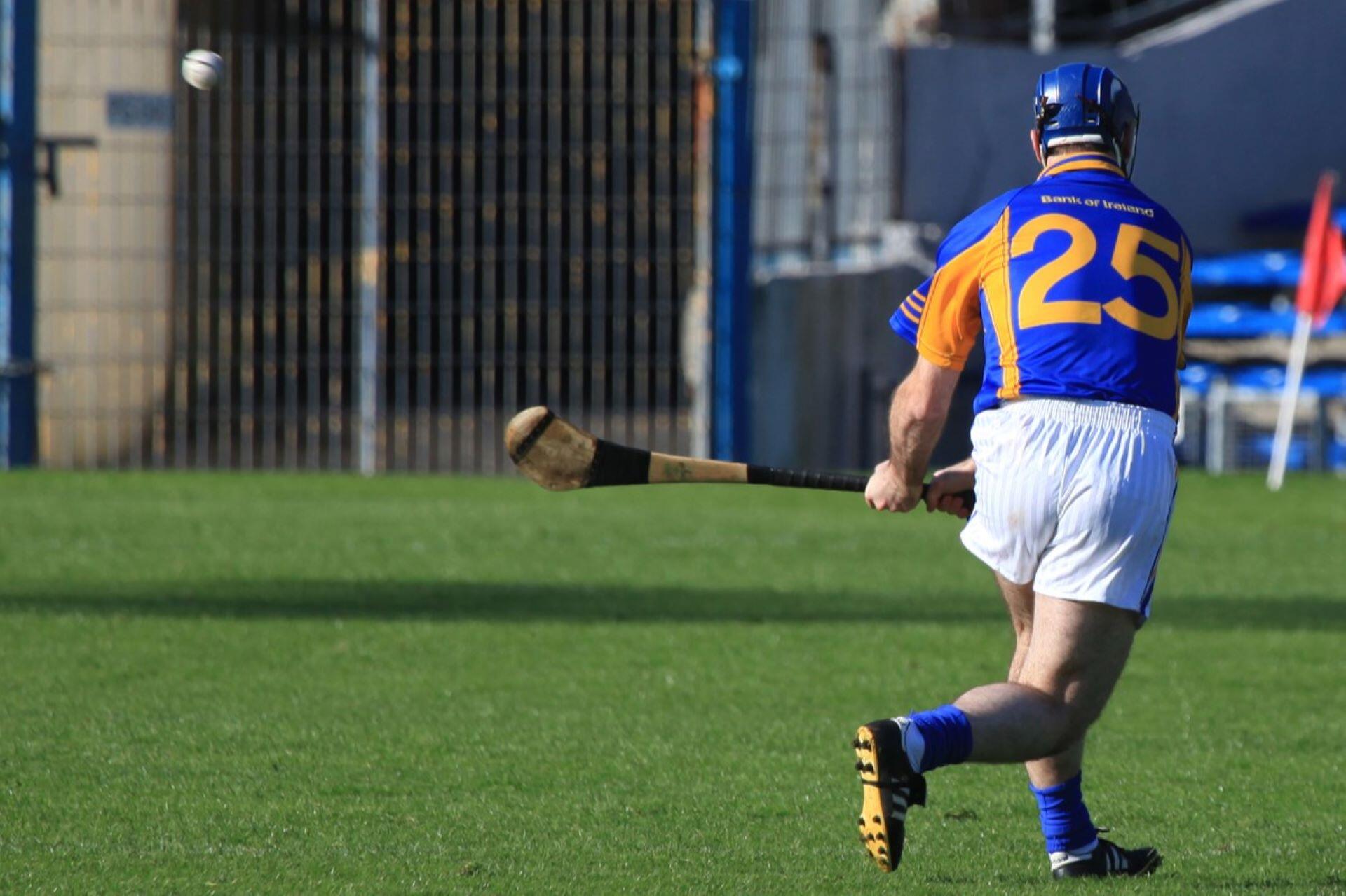 Hurler wearing a blue and gold number 25 jersey striking a sliotar on a grass pitch.