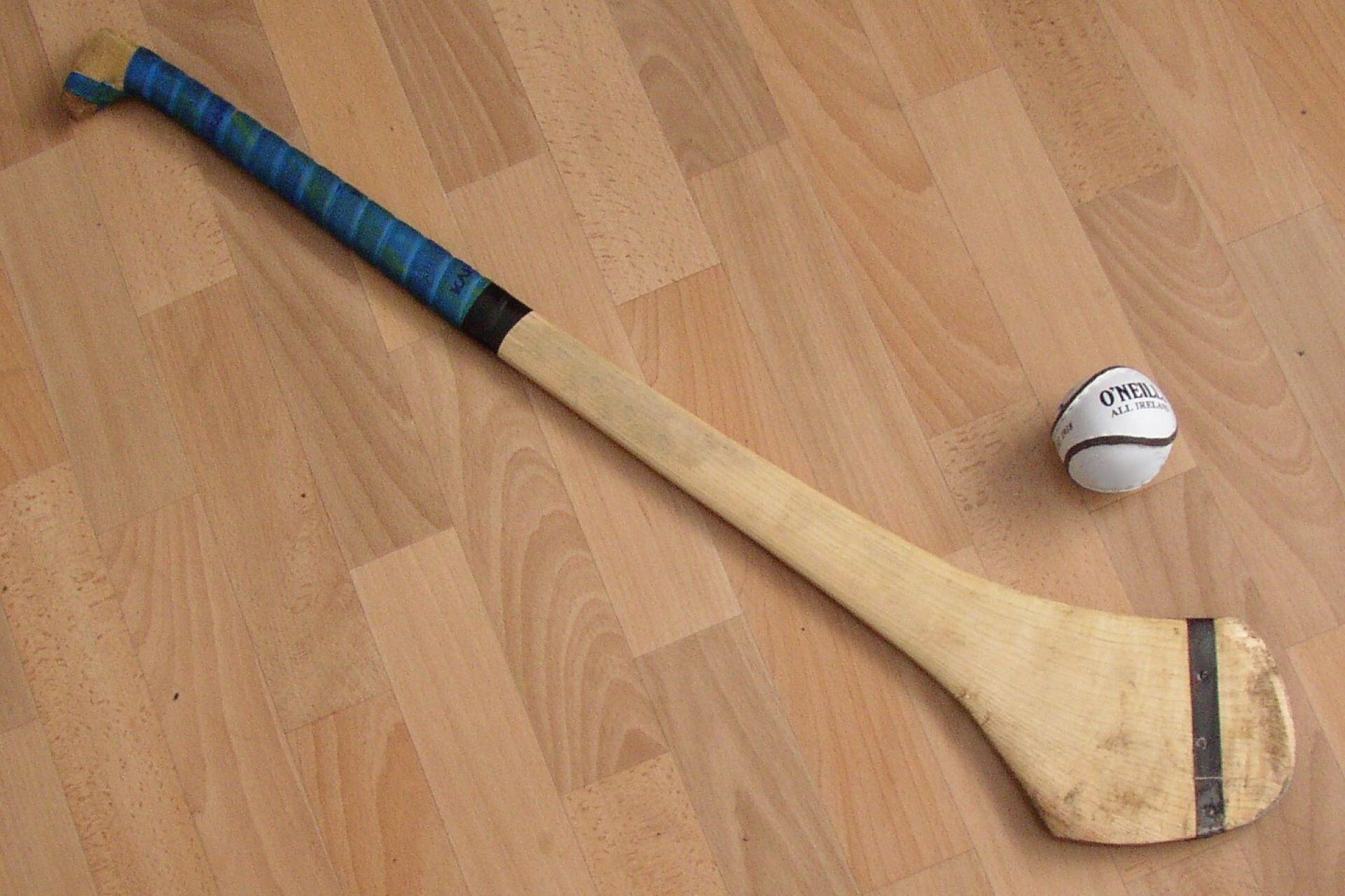 Wooden hurley with a blue grip beside a white sliotar on a wooden floor.