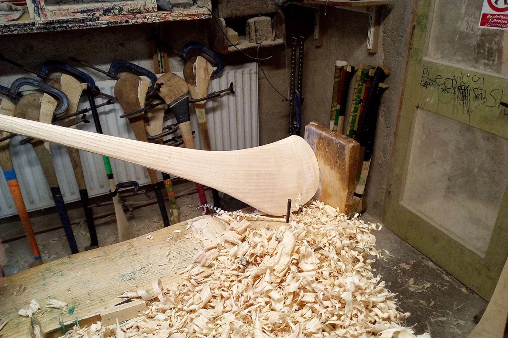 Unfinished wooden hurley clamped to a workbench above wood shavings, with tools and other hurleys behind it.