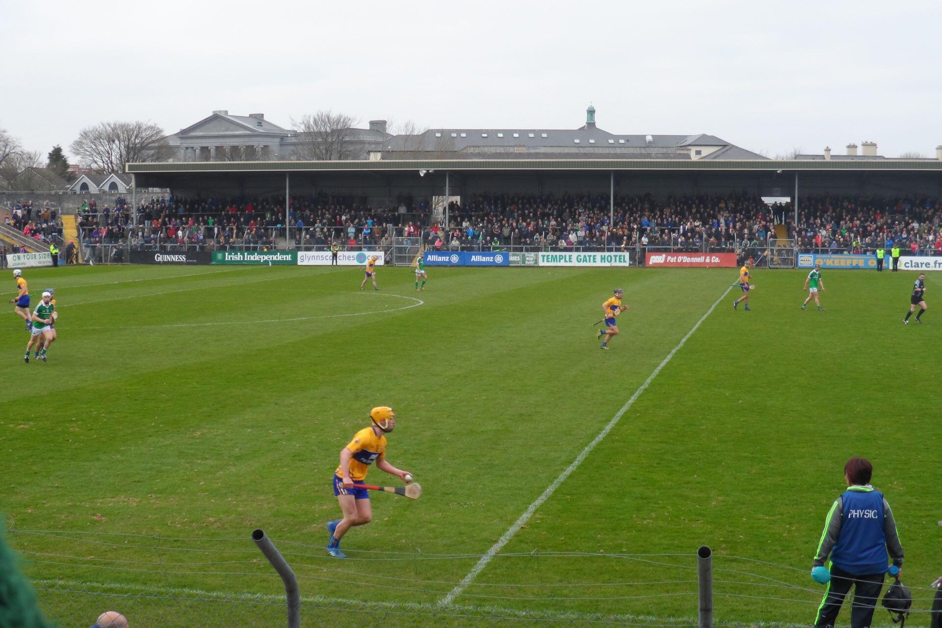 Hurling players in yellow-and-blue and green-and-white kits compete on a grass pitch in front of a crowded stand.