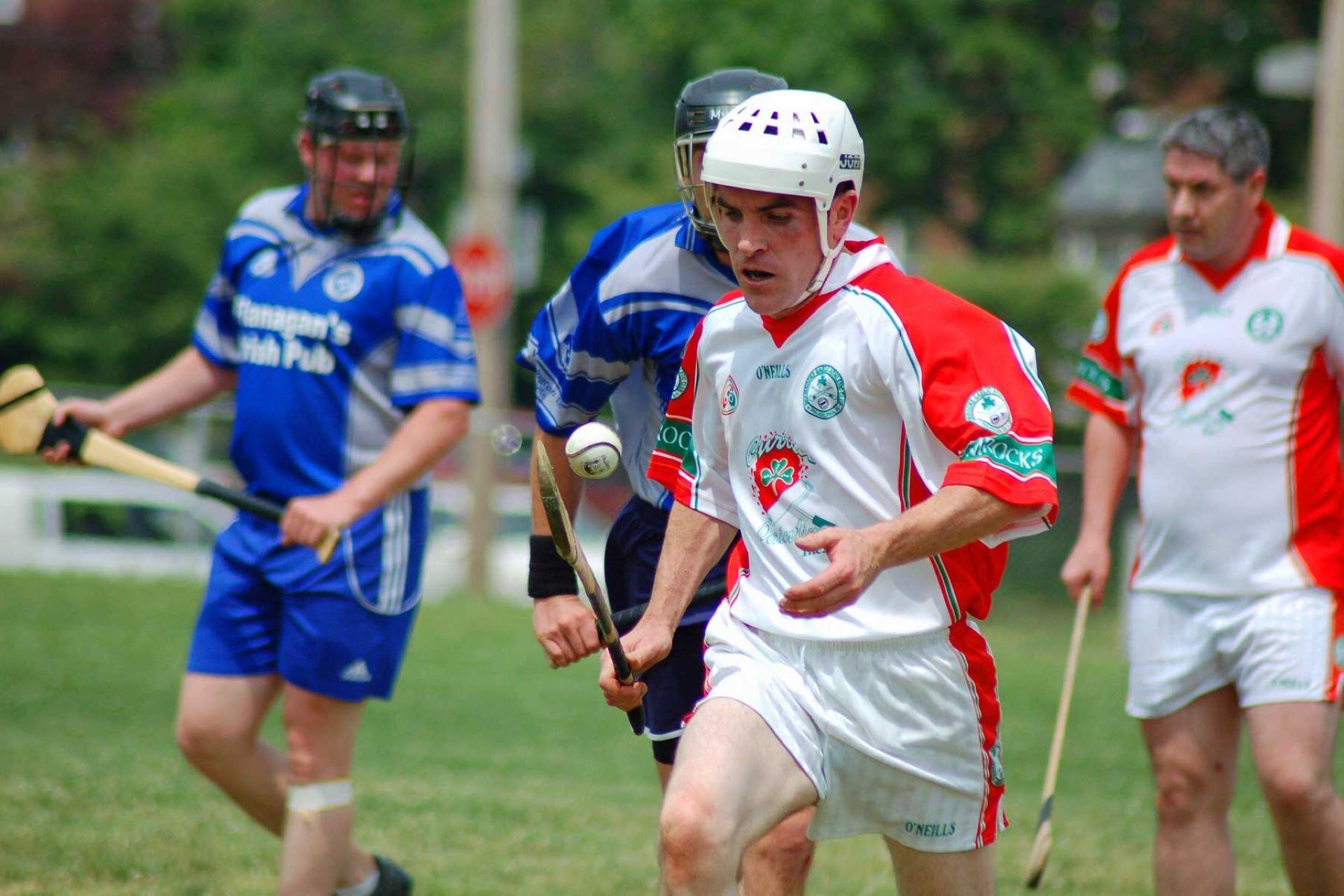 Helmeted hurling player in a white and red kit runs with a sliotar beside players in blue.