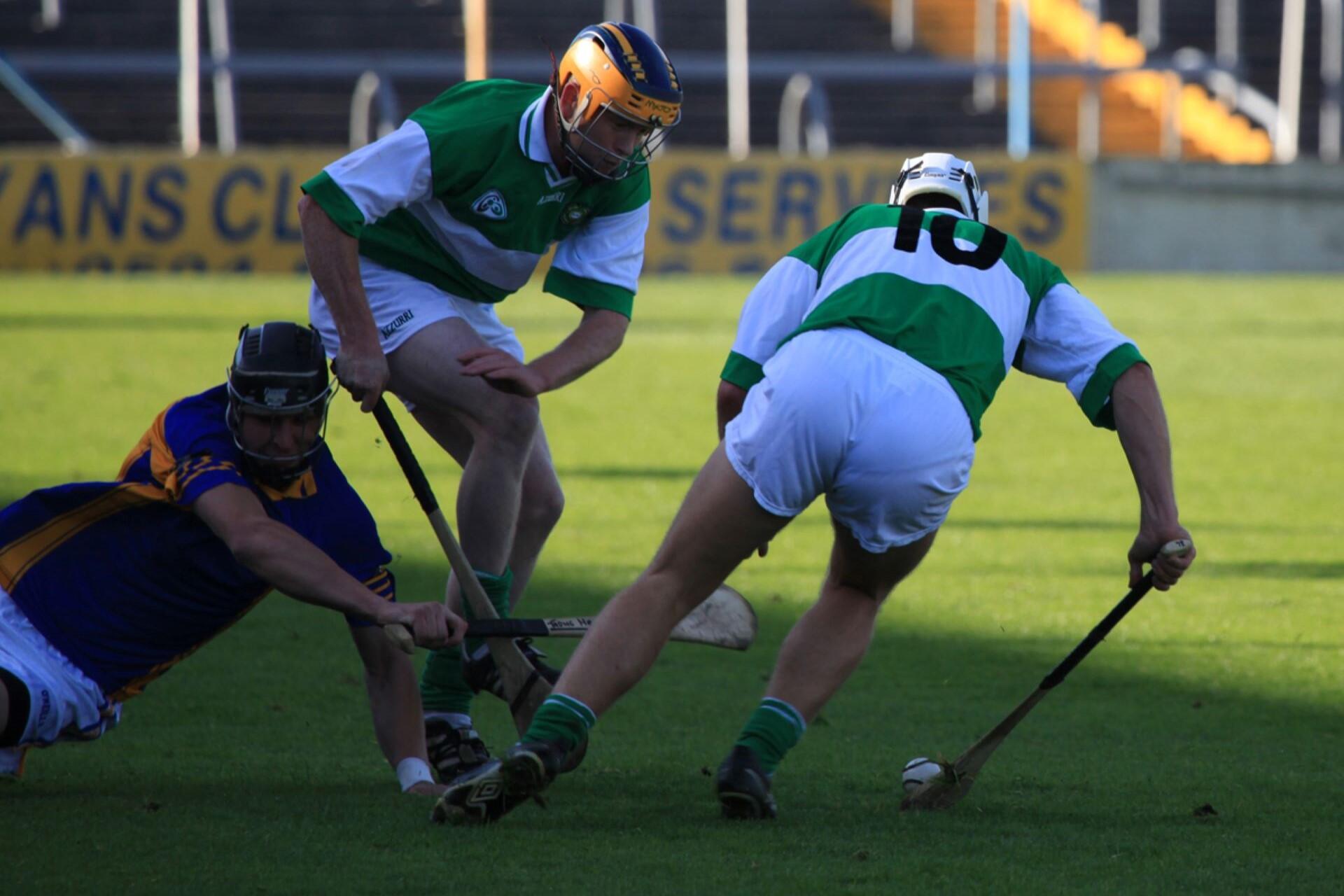 Three helmeted hurling players contesting a sliotar on a grass pitch.