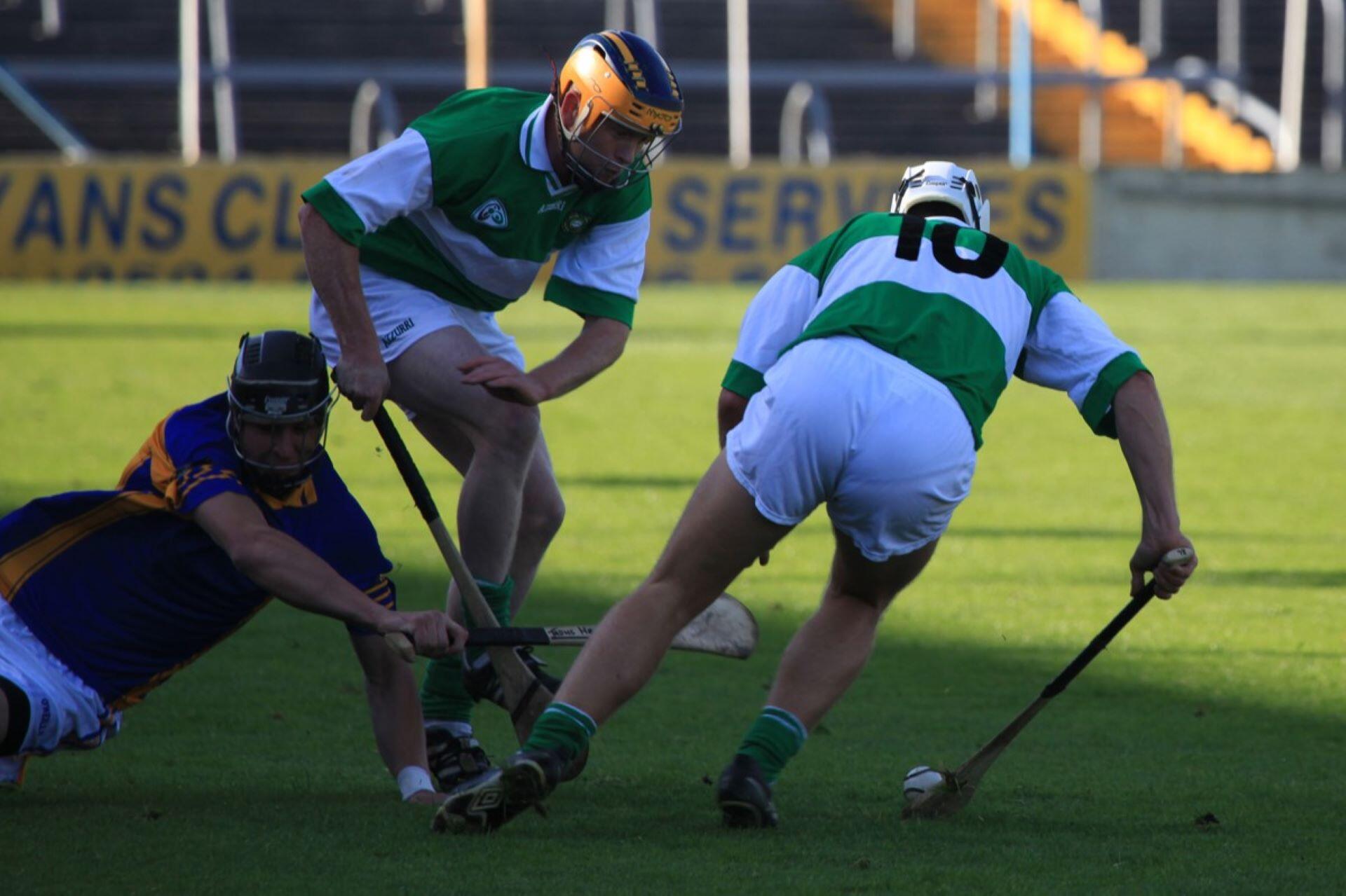 Three helmeted hurling players contest a sliotar on the grass with their hurleys.
