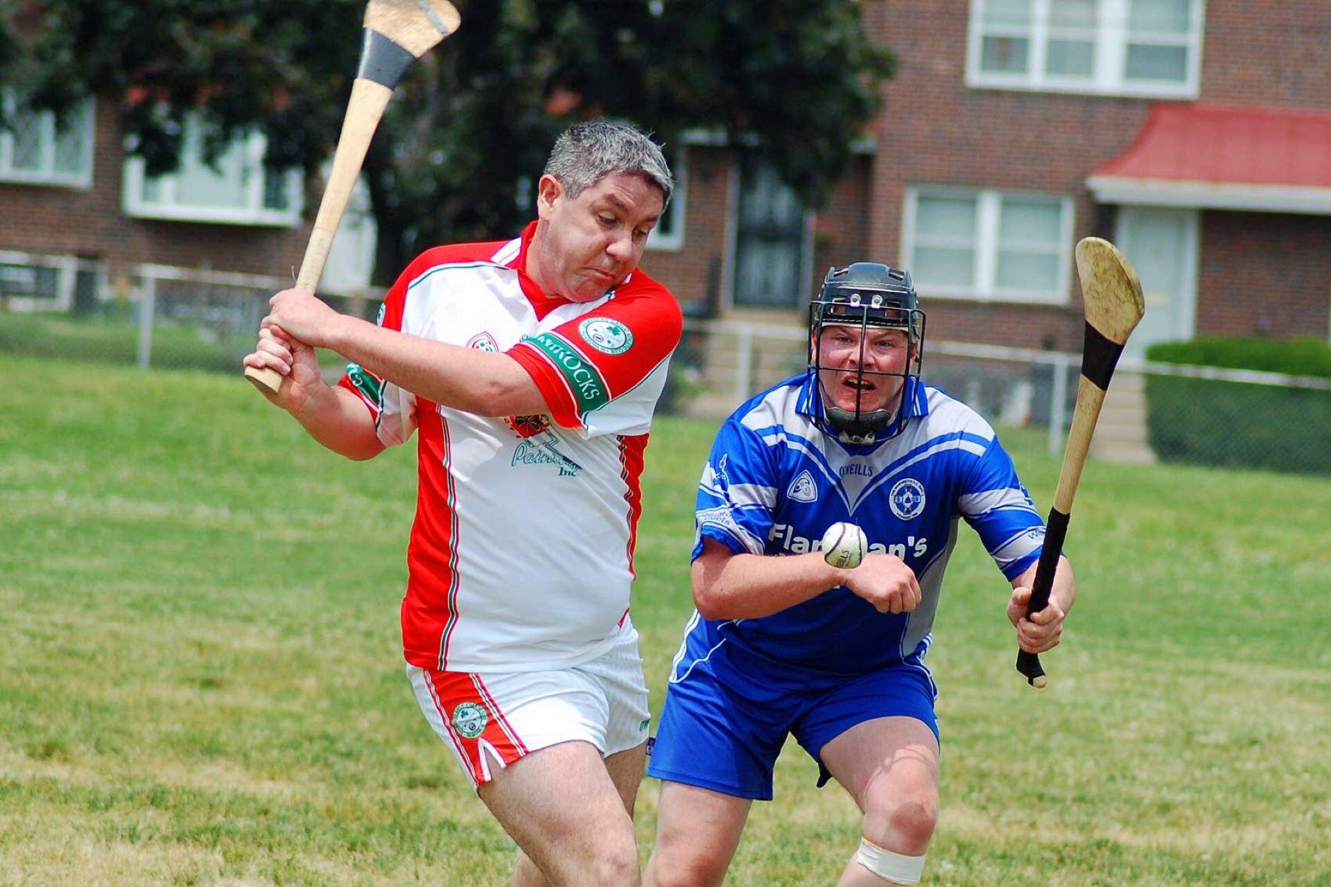 Two hurling players in red-and-white and blue kits compete as one swings a hurley.