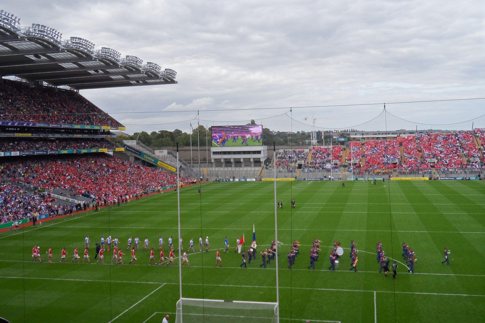Hurling teams and a marching band parade across a packed stadium pitch before a match.